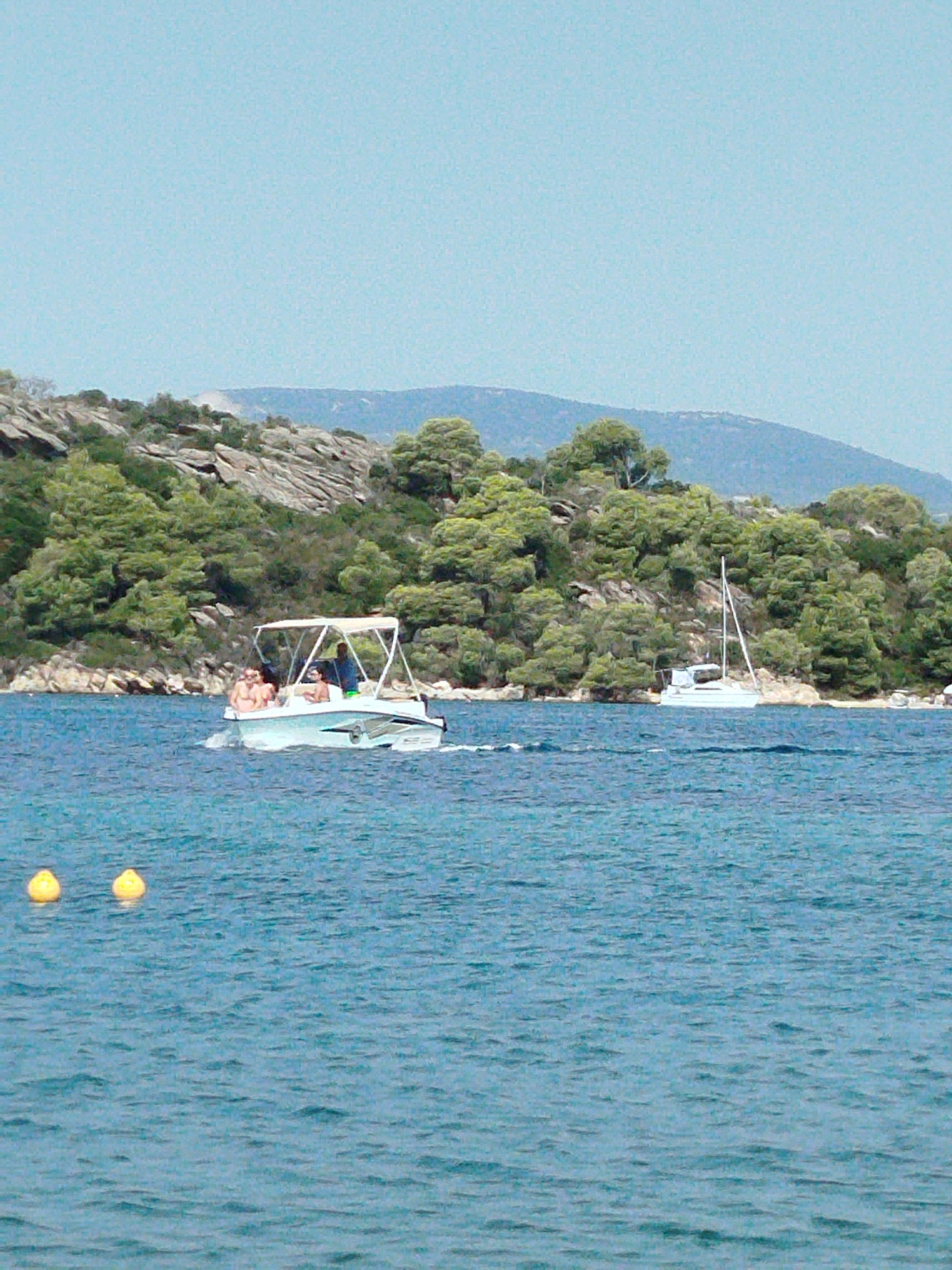 The image depicts a coastal scene with two boats on the water. In the foreground, a small white motorboat with a canopy is moving from left to right, carrying at least three people. Two individuals are visible in the front of the boat, appearing to be sunbathing, while another person stands at the helm. To the right and further in the background, a white sailboat with its mast upright is stationary in the water. The boats are in a body of water that appears to be the sea, with gentle ripples on its surface. Two yellow buoys are visible in the lower left corner of the frame, floating in the water. The background features a rocky, tree-covered hillside with a range of mountains extending into the distance under a clear, light blue sky. The vegetation on the hillside consists of dense green trees and shrubs. The scene is set in a sunny, outdoor environment, likely a popular tourist or recreational area. The location is identified as Sithonia Municipal Unit, Greece.