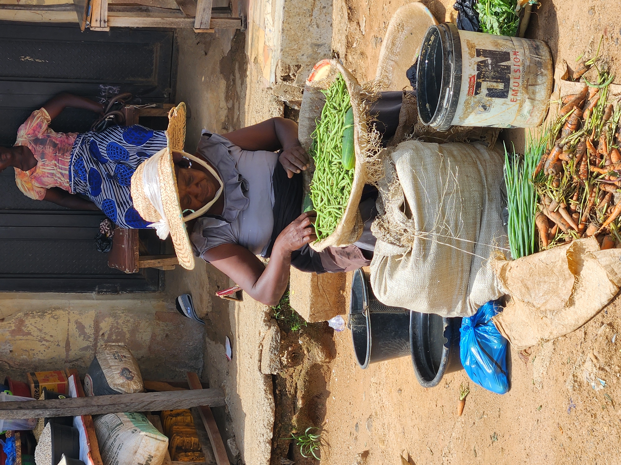 The image shows two women at a roadside market in an unidentified city. The vendor in the foreground wears a straw hat and is sorting green beans in a large, fiber basket. Various other vegetables, including carrots and green onions, are displayed for sale. A white bucket labeled with "Emulsion" rests near the produce. A blue plastic bag sits on the ground. The second woman is resting on a stool near a door, wearing a patterned dress. The environment appears to be a dusty, outdoor area.