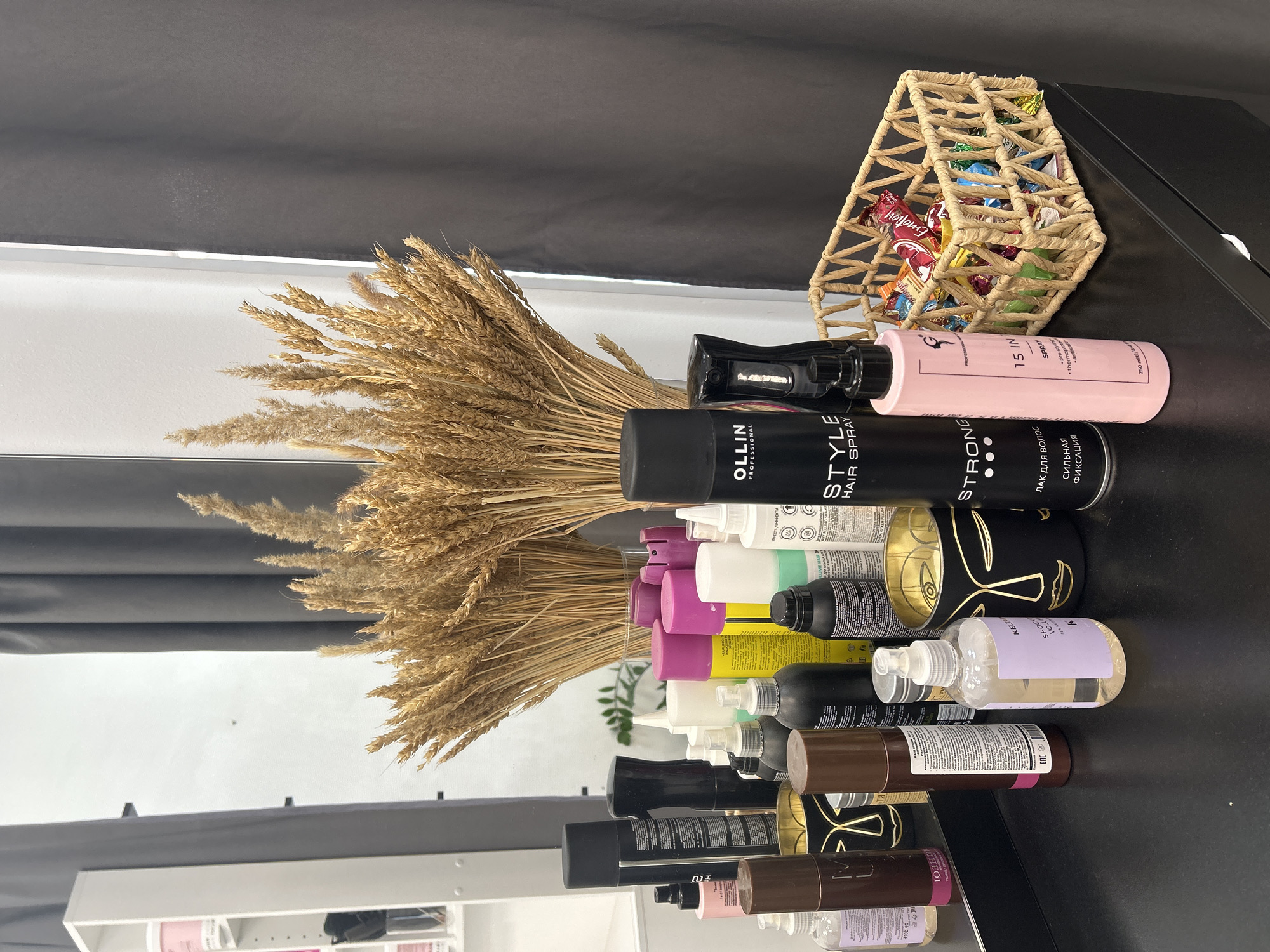 This image depicts an interior scene, likely a beauty salon or a dressing area, in Chaikovsky, Russia. The foreground features a black countertop upon which numerous objects are arranged. These objects include a collection of hair care products in various bottle types (aerosol cans, spray bottles, pump bottles) and colors (black, pink, brown, yellow, white, blue, green). Notably, an Ollin Professional Style Hair Spray can is visible, along with a pink bottle labeled "15 IN 1 Spray" and a clear bottle with a purple label for "KELPSEA Shocking Volume." Two black decorative cylindrical containers with gold abstract face designs are also on the counter. To the right, a rectangular, light-brown woven basket is filled with an assortment of individually wrapped candies.
In the midground, positioned behind the products and reflected in a large mirror, is a substantial bouquet of dried golden wheat stalks, contained within a clear glass vase. The mirror reflects a white wall and a white shelving unit containing some items, visible on the left side of the reflected image. Dark grey curtains frame the right side of the scene, both directly visible and reflected in the mirror. No individuals or explicit actions are present within the frame.