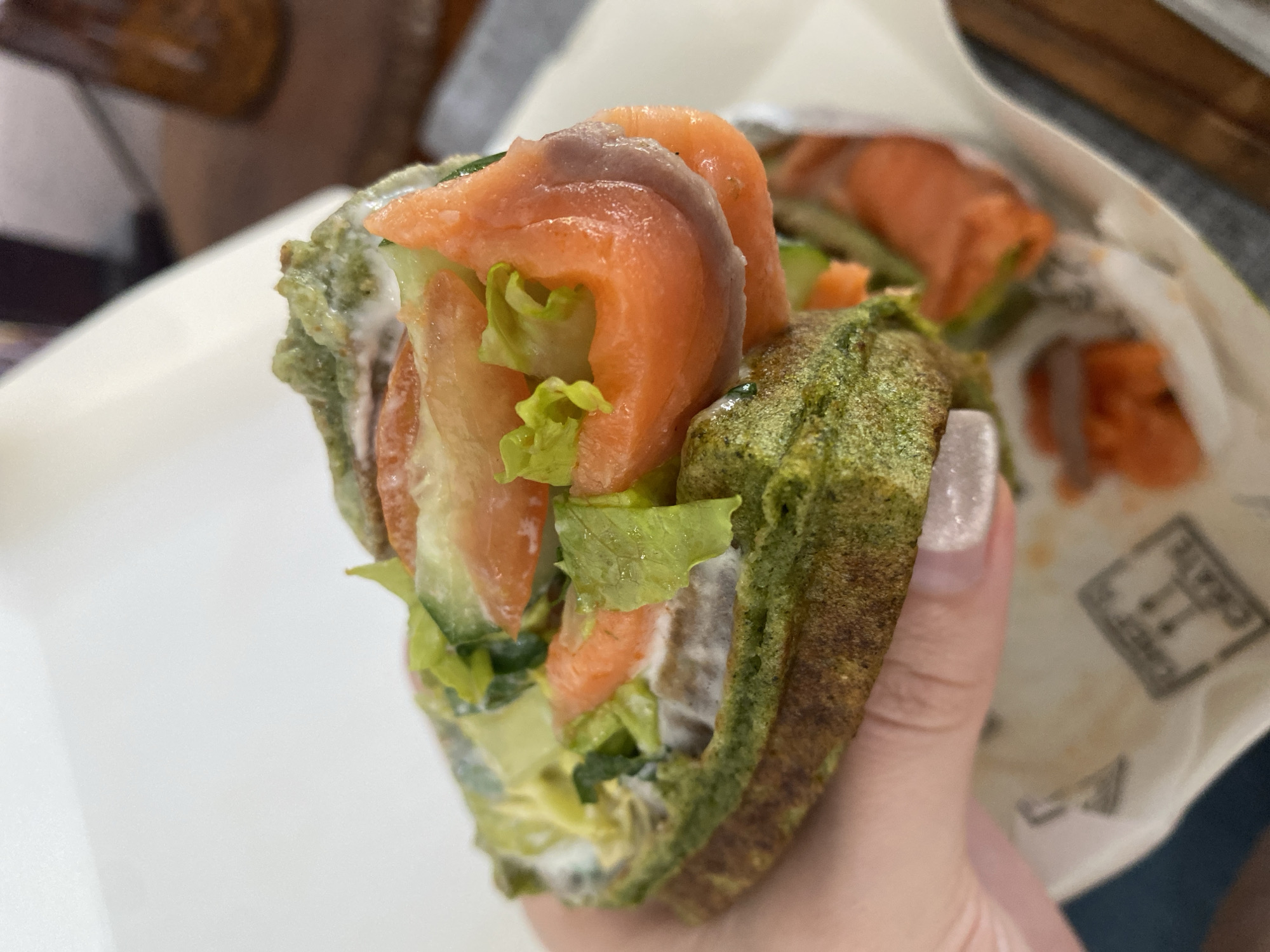 A hand holds a green-colored sandwich filled with salmon, lettuce, cucumber, and a creamy white spread. The sandwich is partially wrapped in white paper with printed text or a logo. The background includes blurred hints of a table, chair, and possibly a bag. This image does not contain enough information to infer the location in Kuta Selatan, Indonesia.