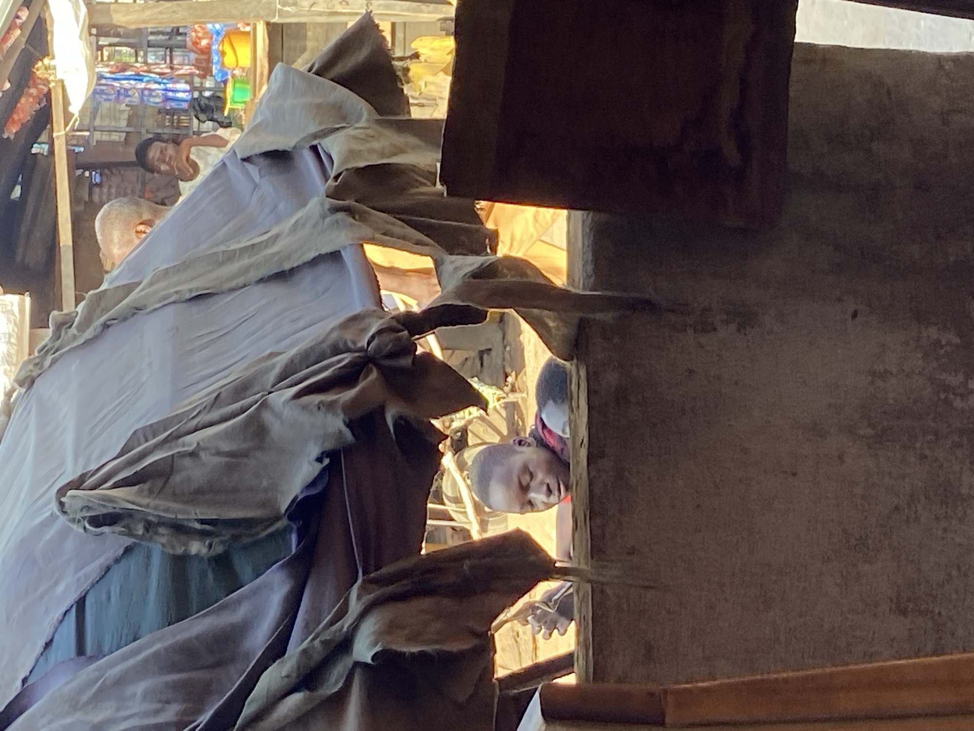 The image captures a scene within a market in Mararaba, Nigeria. In the foreground, a young individual, likely a boy, is peering over a rough, textured wall. He has short hair and appears to be wearing a red garment. Behind him, a large, draped fabric, possibly part of a stall or shelter, hangs down, obscuring much of the background. To the right of the young boy, another person's head, wearing a white top, is visible, looking towards the viewer. Further in the background, there are glimpses of market stalls with various items, including colorful buckets and what appears to be produce. The overall environment suggests a bustling outdoor market setting.