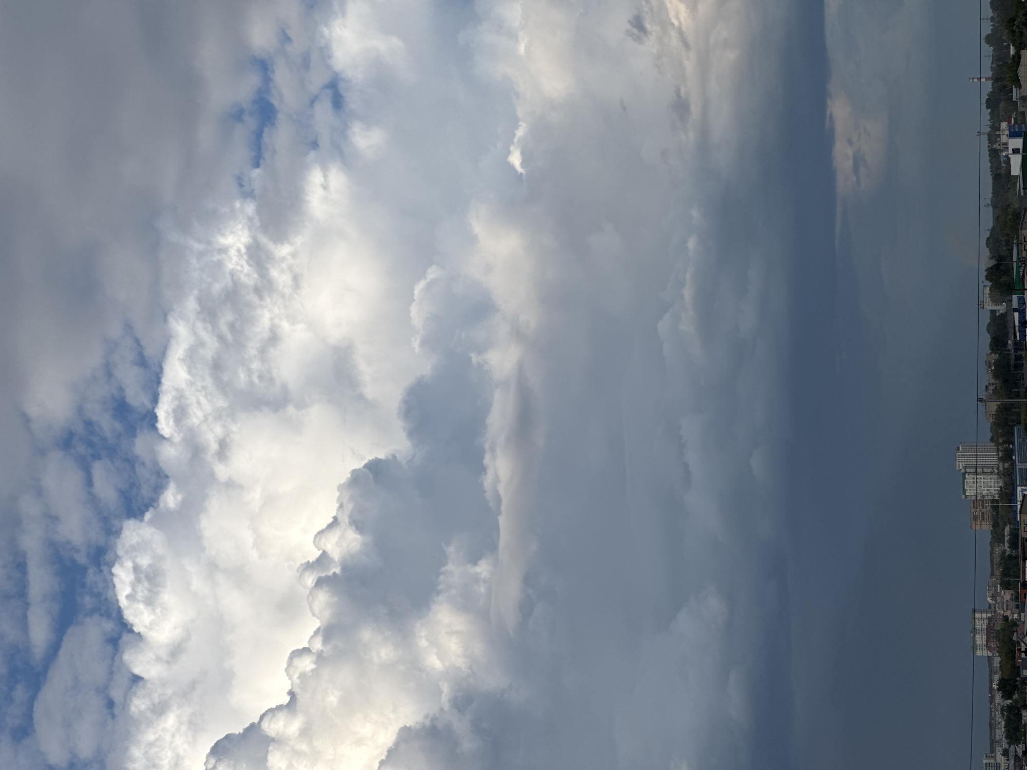 The image depicts a dramatic sky over a cityscape, likely in Novosibirsk, Russia. The upper portion of the frame is dominated by a tumultuous sky filled with a variety of clouds. Bright, white cumulus clouds are prominent in the top left and center, suggesting sunlight breaking through. These are contrasted by darker, heavier grey clouds that fill the middle and lower portions of the sky, hinting at an impending storm or recent rainfall.
Below the dense clouds, a distant cityscape is visible. Numerous buildings of varying heights and architectural styles are scattered across the horizon. Some appear to be residential apartment blocks, while others suggest industrial or commercial structures. Green trees and vegetation are interspersed among the buildings, indicating a semi-urban or suburban environment. A thin, dark horizontal line, likely a power line or wire, stretches across the lower part of the image, suggesting an elevated viewpoint. The overall impression is one of atmospheric dynamism, with the powerful sky creating a stark backdrop for the human settlement below.