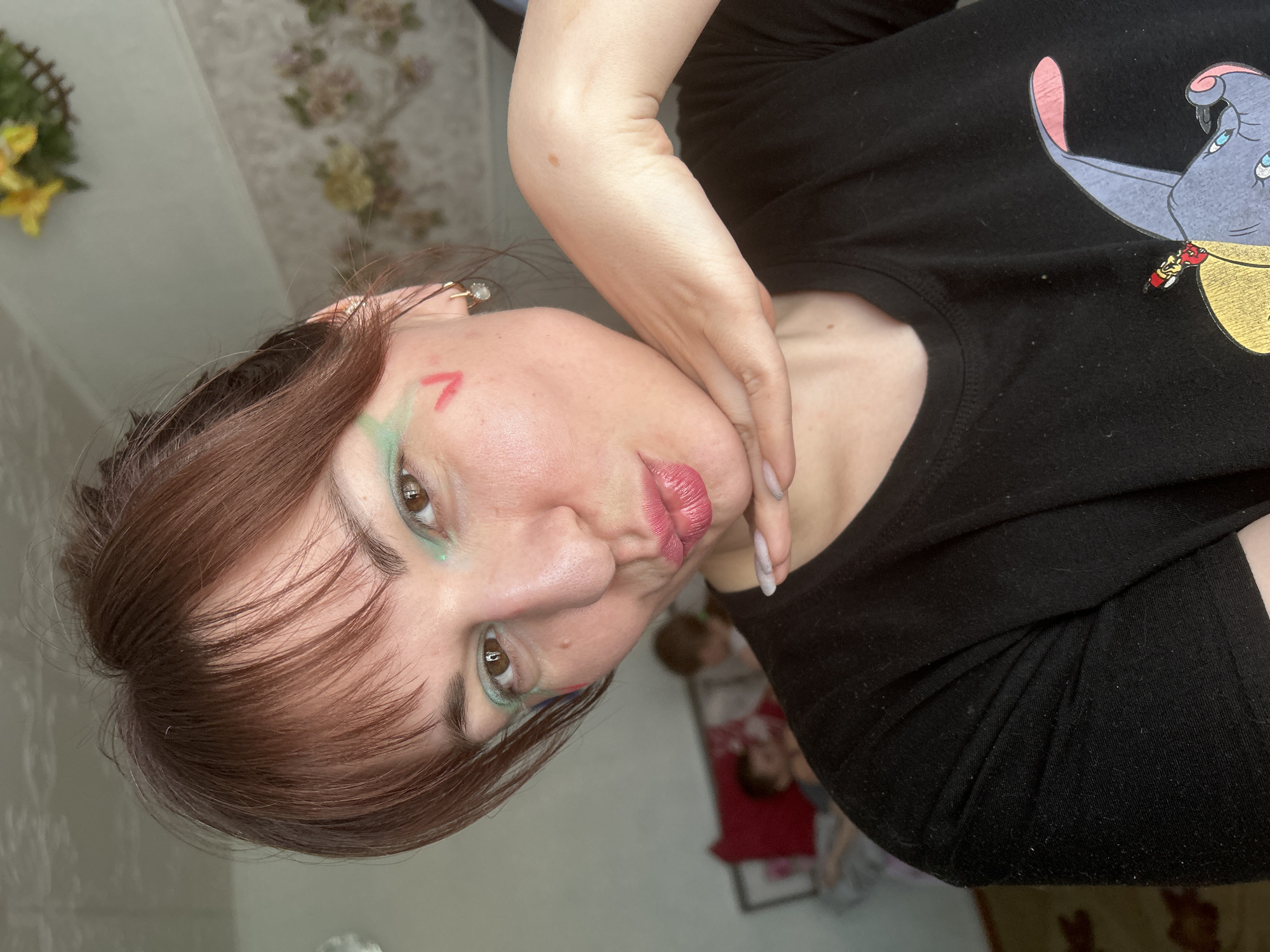 A female subject is depicted in a medium-shot selfie, posing indoors in what appears to be a residential setting within the city of Фоки, Russia. She has brown hair with bangs, fair skin, and is looking directly at the camera with a puckered, kissing expression. Her eyes are accentuated with green eyeliner/eyeshadow. A red inverted 'V' shape is painted on her right cheek, and faint red and blue markings are visible beneath her left eye. She wears small, light-colored hoop earrings. Her right hand, with light-colored manicured nails, rests under her chin. She is wearing a black t-shirt featuring a graphic print of Dumbo the elephant and Timothy Mouse on the chest. In the blurred background, two individuals, likely children, are partially visible on a bed with a red pillow. The room features light-colored walls, with floral patterned wallpaper visible on the right and a plain wall on the left. A decorative element with yellow artificial flowers is visible in the upper right background. A patterned rug is partially visible on the floor in the lower left.