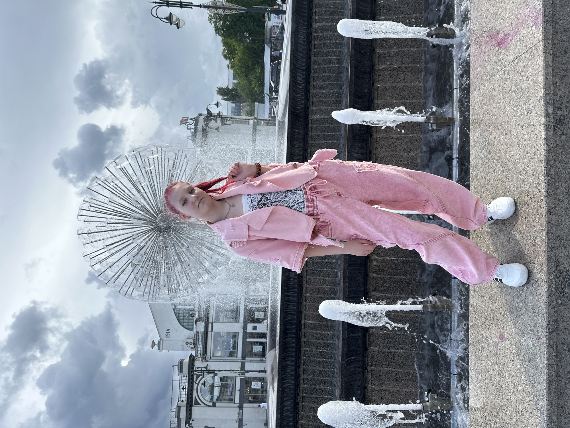 A full shot features a young person with reddish hair wearing a pink denim outfit and white athletic shoes, standing on the side of a fountain in Saratov, Russia. The person is positioned adjacent to the fountain's edge, which features small water jets. Behind the person, a large, spherical fountain is visible, along with a building marked with "1914". Above, a cloudy sky is present. The image appears to be taken at street level.
