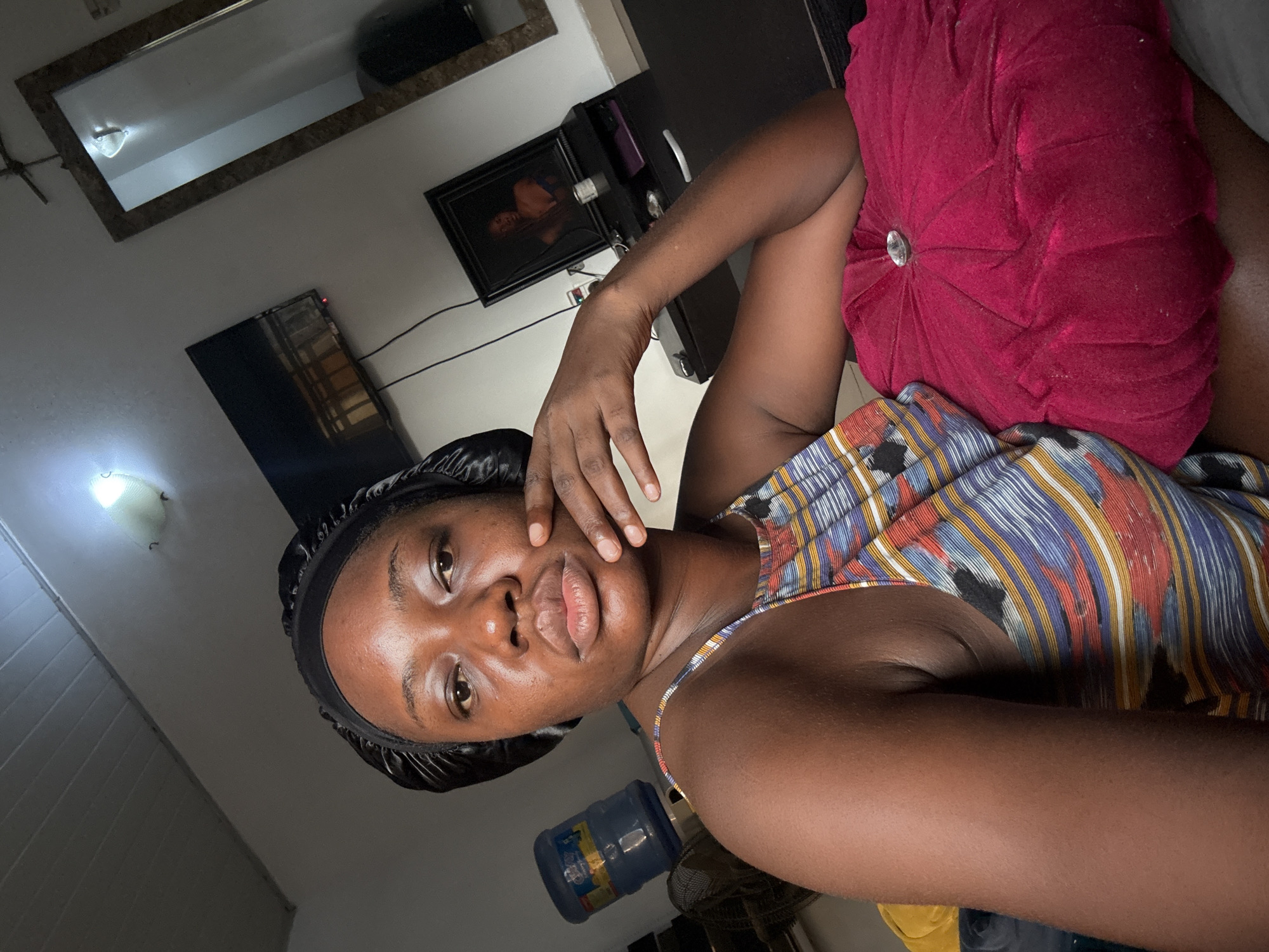 A person with brown skin and wearing a black hair covering is pictured indoors, possibly in a bedroom. They are wearing a colorful patterned top and have a round, deep red cushion placed beside them. In the background, a wall with a mirror, a framed image, and a television is visible. A water bottle is near the person. The location is indeterminate.