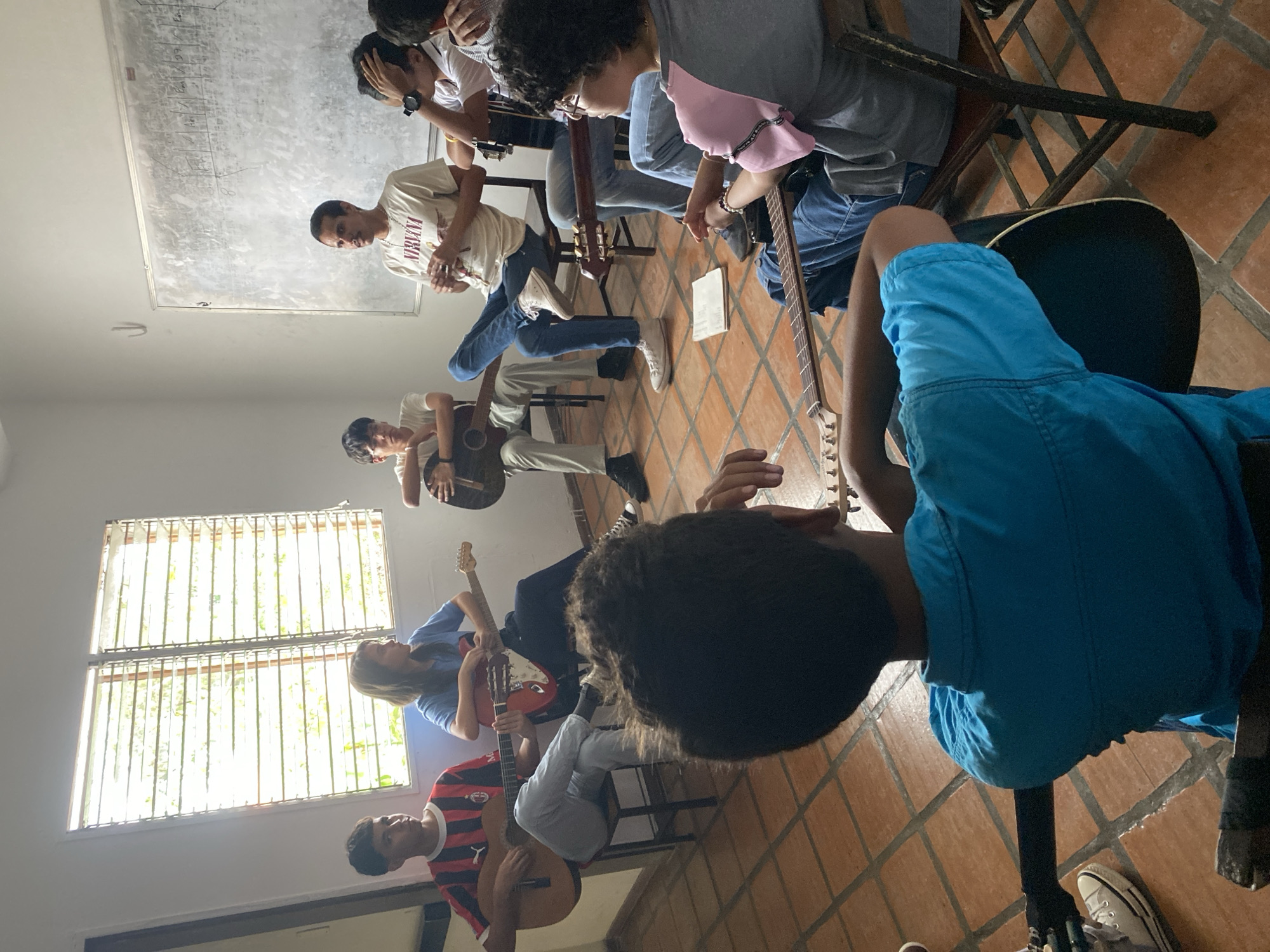 The scene depicts a group of approximately 8-10 young men engaged in a guitar lesson inside a room in Tinaquillo, Venezuela. The room features a tile floor, white walls, and a window with bars. A whiteboard with writing is visible on the wall. Each person is seated and holding a guitar, appearing to be in a lesson or practice session. One young man wears a "Nirvana" T-shirt. The room seems to be well-lit.