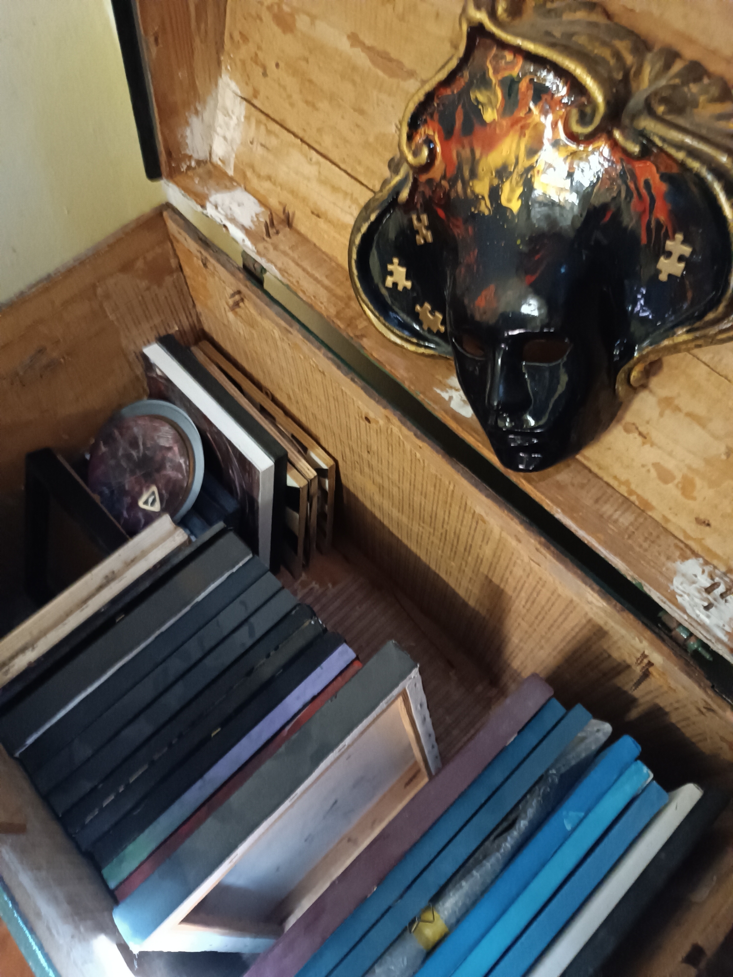 A wooden chest is open, revealing an assortment of objects inside. A black and gold mask with puzzle piece details rests on the open lid. Inside the chest, there is a stack of framed objects or canvases, many with black edges and some with other colors like blue, purple, and green. There is also a round object among the frames. The photograph does not contain enough information to confirm it was taken in Viadana, Italy.