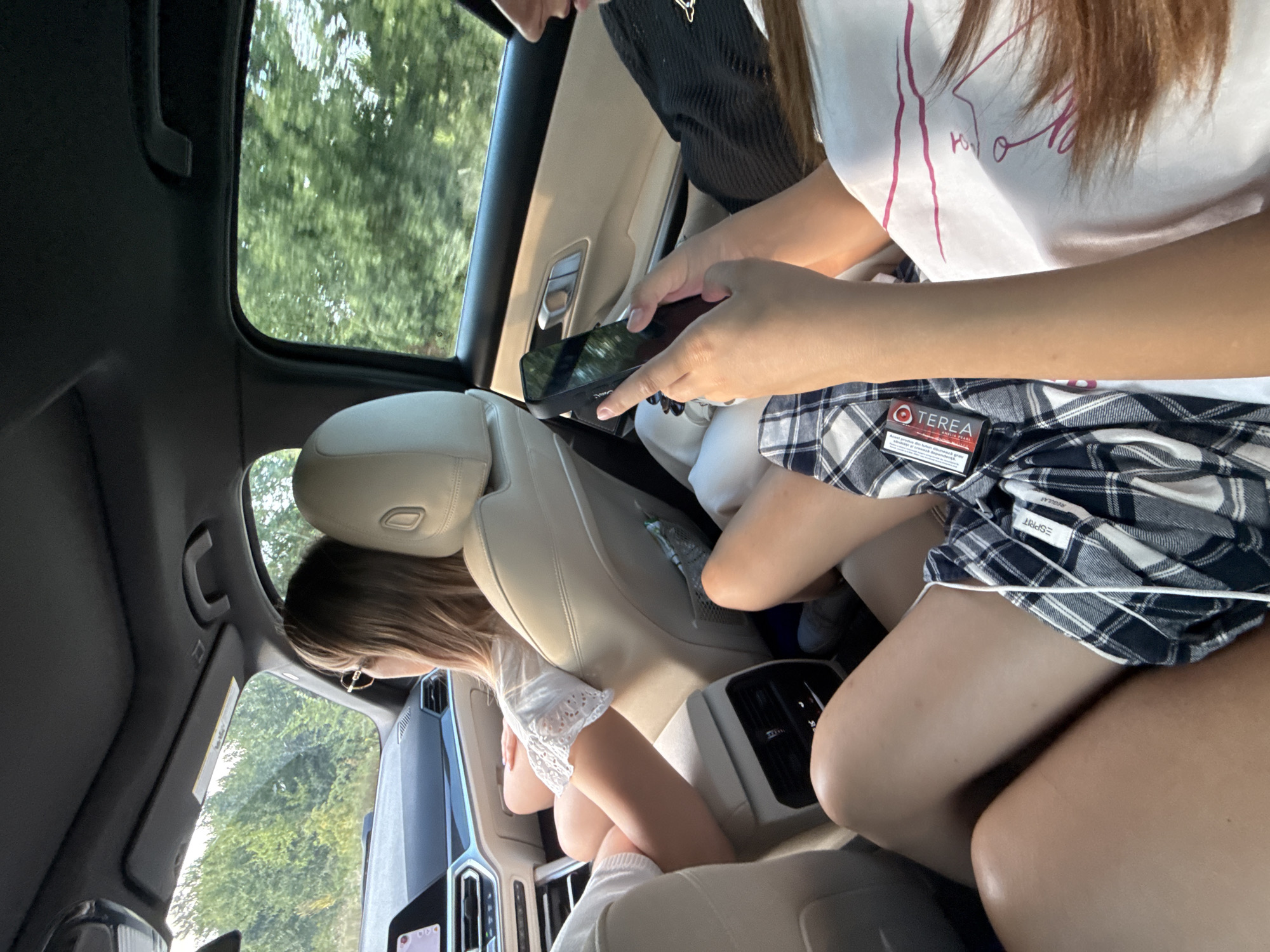 The image depicts the interior of a car traveling on a road, with lush green trees visible through the windows, suggesting a rural or suburban setting, potentially on the outskirts of Onești, Romania. Three young females are seated in the rear of the vehicle. The person in the foreground on the right is wearing a white t-shirt with pink lines and has long brown hair. To their left, another female is wearing a black top and is looking down at a smartphone she is holding. In front of them, a third female wearing a white top with lace trim and sunglasses is seated, looking forward. The interior of the car features light-colored leather seats, a black headliner, and a dashboard with a modern infotainment system. The overall scene captures a moment of travel, with a focus on the passengers and their activities within the vehicle.