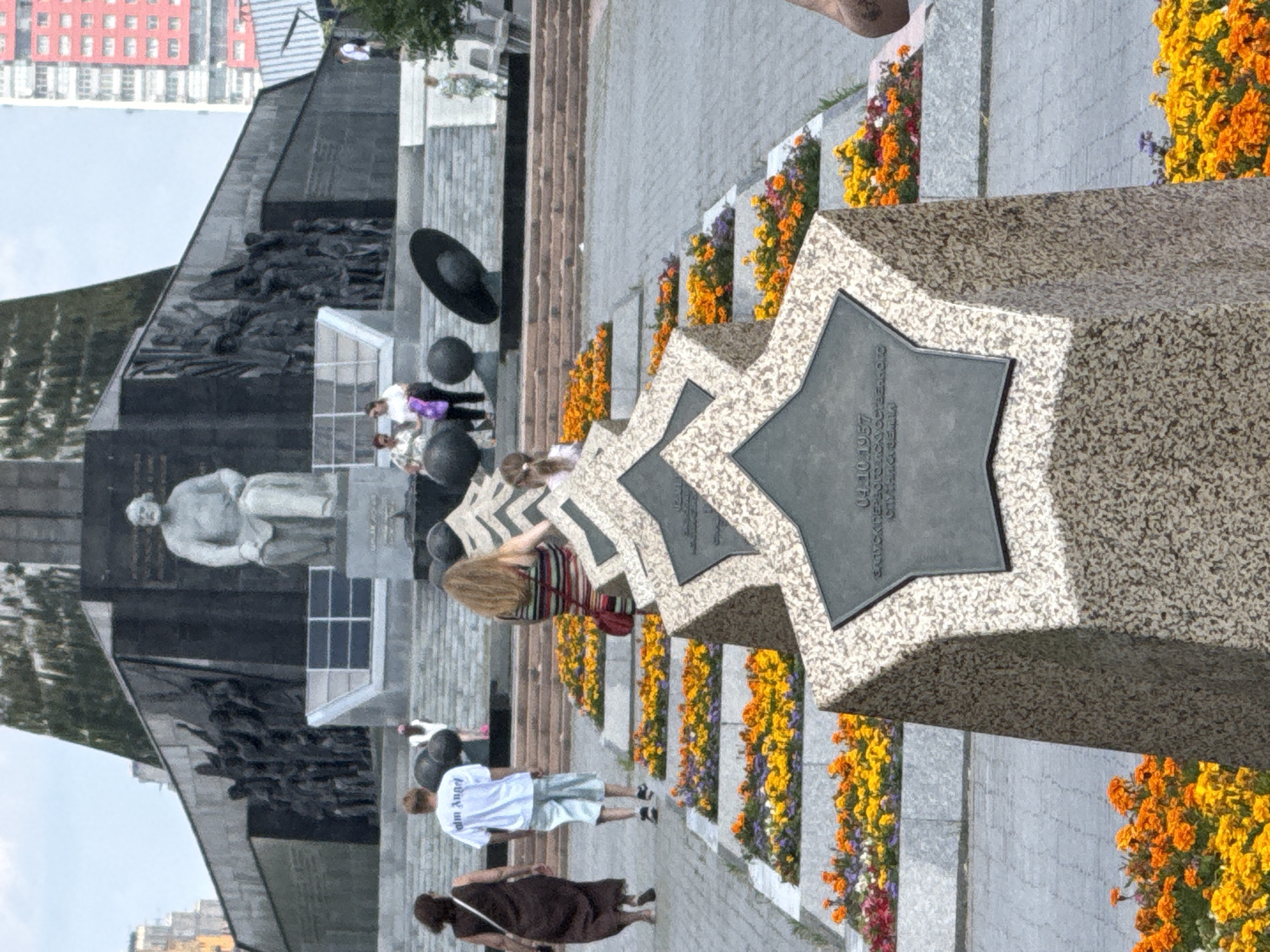A memorial site in Moscow, Russia, is depicted with a large granite monument featuring a star-shaped plaque inscribed with text. The monument is surrounded by a paved walkway lined with vibrant flower beds in shades of orange, yellow, and purple. In the background, a monumental sculpture of a seated figure stands against a dark, imposing structure adorned with relief carvings of people.
Several individuals are present at the site. A person in a white t-shirt and light-colored shorts stands facing away from the camera, while another individual in a dark dress walks by. Further in the background, a few people are gathered near the monumental sculpture, some appearing to be observing the artwork. The overall scene suggests a public space, possibly a park or a memorial ground, where people are visiting.
The time of day appears to be daytime, with bright sunlight casting shadows, indicating fair weather. The architecture and the style of the sculptures suggest a historical or solemn context. Visible text on the star-shaped plaque includes "10.1957" and what appears to be Russian characters, possibly indicating a date or dedication.