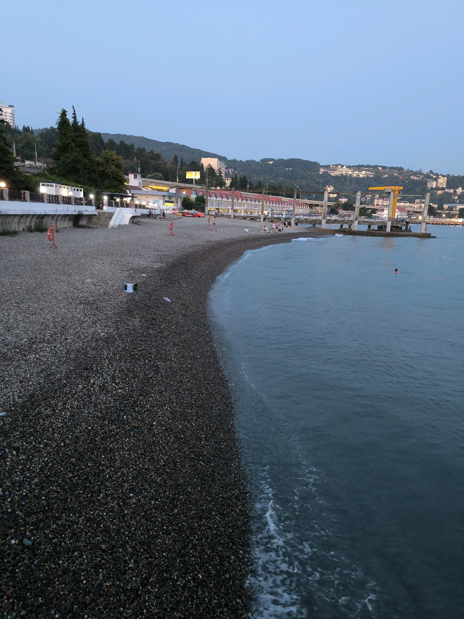 A pebble beach lines the coast of Sochi, Russia, at dusk. The sea is a calm, dark blue with gentle waves lapping the shore. In the distance, a pier with a yellow crane juts out into the water, and a cluster of buildings, some with illuminated windows, dot the hillside behind the beach. Several people are visible on the beach, some walking and others sitting. One person is swimming in the sea. The sky is a pale, twilight blue.
