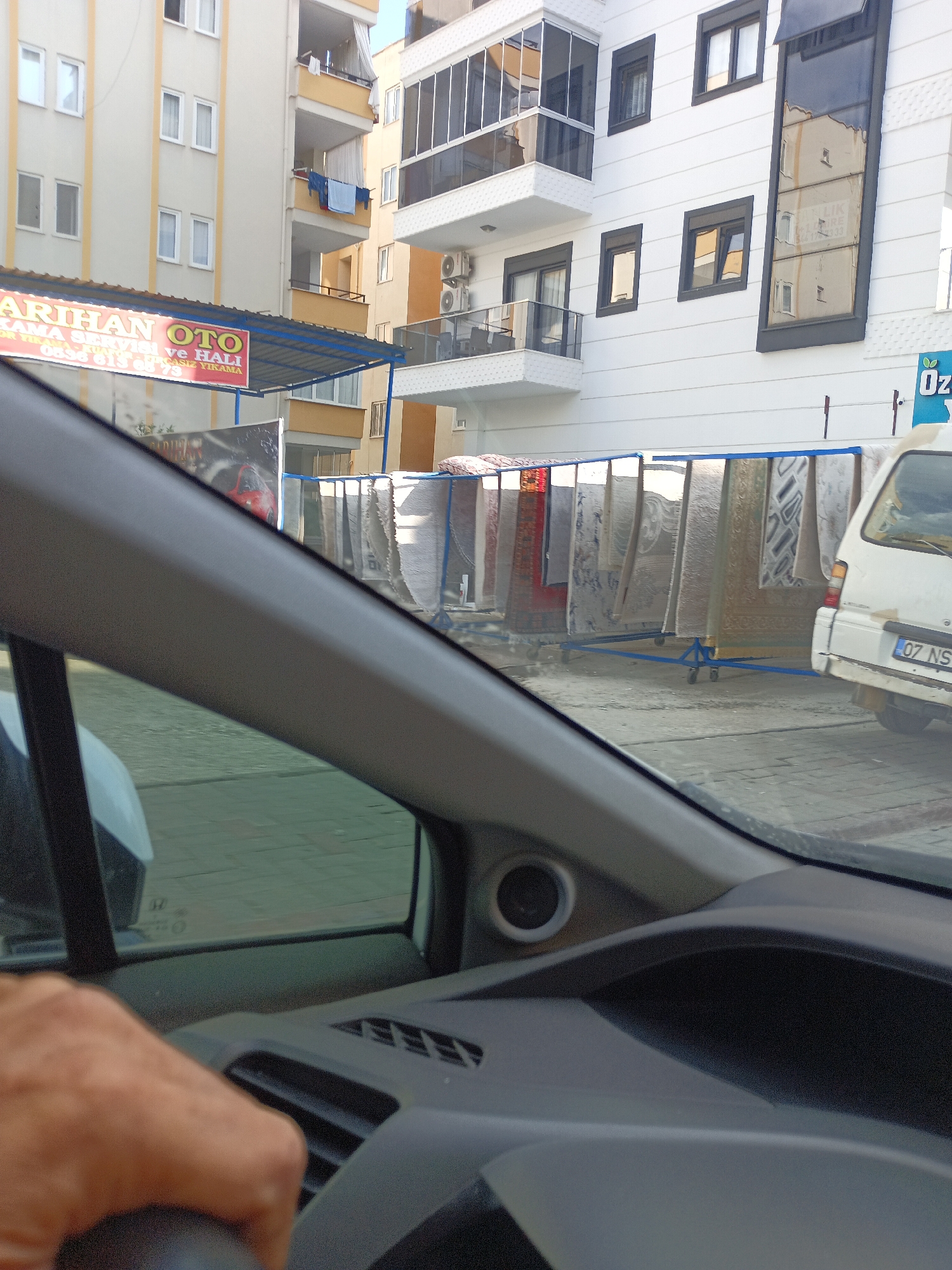 The image shows a street scene in Alanya, Turkey, viewed from inside a vehicle. A building with multiple floors is visible on the right, featuring modern white facade with dark window frames and balconies. To the left of this building, there is another apartment building with a cream-colored facade. In front of the buildings, a display of carpets is set up on blue racks, with some carpets hanging and others rolled. A white van with a license plate starting with "07 NS" is parked to the right of the carpets. A red car is partially visible in an advertisement banner behind the carpets. The foreground shows the interior of a car, including a hand on the steering wheel and part of the dashboard. The overall environment appears to be an urban street, possibly a business district or residential area.