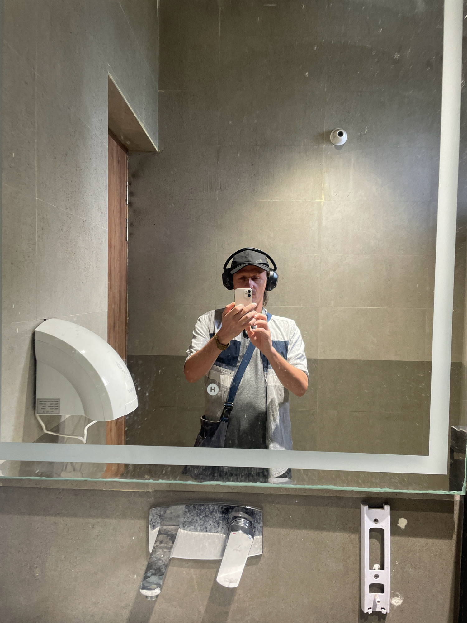 A man wearing a black baseball cap and headphones is taking a selfie in a restroom mirror. He is holding a white smartphone in his right hand and is wearing a white t-shirt with blue accents and a blue strap bag over his left shoulder. The restroom has gray tiled walls and a large mirror. To the left of the man, a white hand dryer is mounted on the wall. In the foreground, a chrome faucet is visible. The scene is consistent with a modern public restroom in Dharamshala, India.