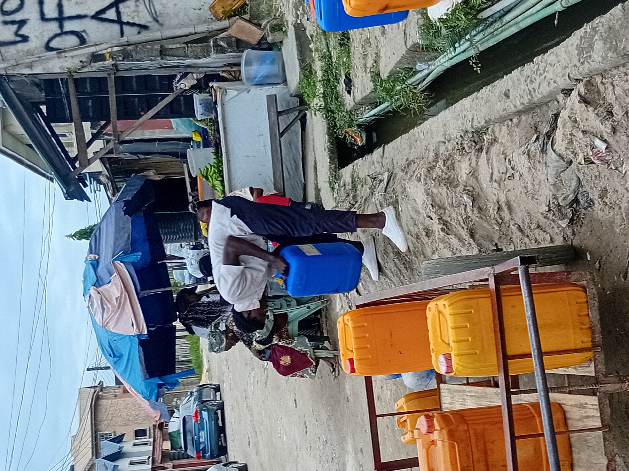 In an outdoor urban environment, possibly a market or roadside stall area, a person in a white shirt and dark pants is bending over, holding a large blue plastic container. The container is upright and appears to be full. The person is wearing white sneakers. In the foreground, a red-railed cart contains several large yellow and orange plastic containers, some with red lids. Behind the person, an older woman in a colorful headscarf and dress sits on a plastic chair. Other people are visible in the background. A blue umbrella is propped up over some of the individuals. To the left, a blue car is parked on a dusty surface. A narrow concrete drainage channel runs along the right side of the image, with green pipes and some vegetation. The background shows buildings and overhead wires. The overall scene suggests a commercial area with goods being transported or sold.