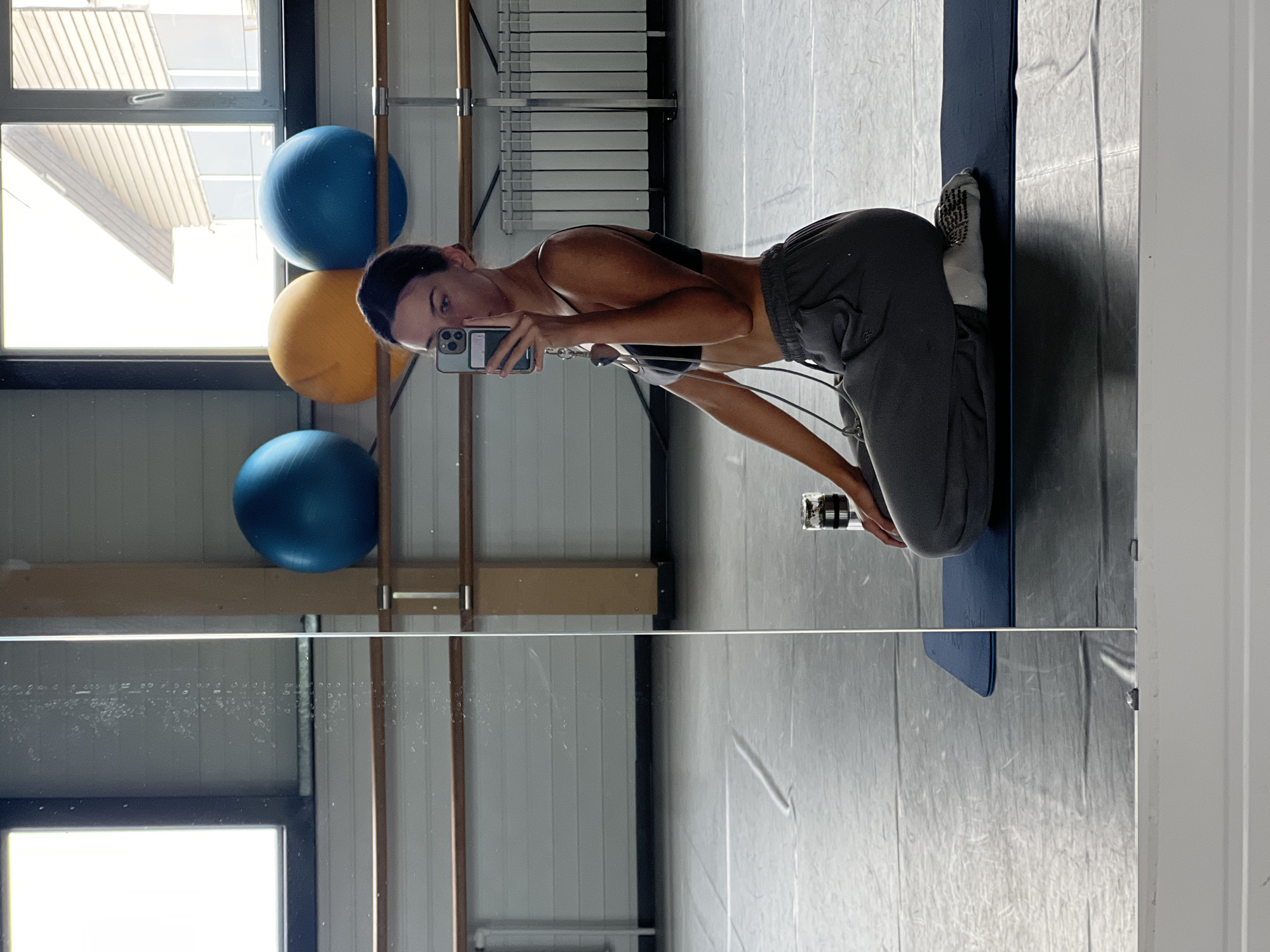 A young woman in athletic attire is taking a selfie in a gym. She is kneeling on a blue yoga mat, wearing a black sports bra and gray sweatpants, with athletic shoes on her feet. She holds a smartphone in her hand, capturing her reflection in a mirrored wall. Behind her, there are three exercise balls—two blue and one yellow—positioned next to a window. Gym equipment, including bars, is visible in the background. The gym features a grey floor. This photograph was likely taken in a gym in a place such as Zima Juzhnaja, Russia due to similar photos from the area.