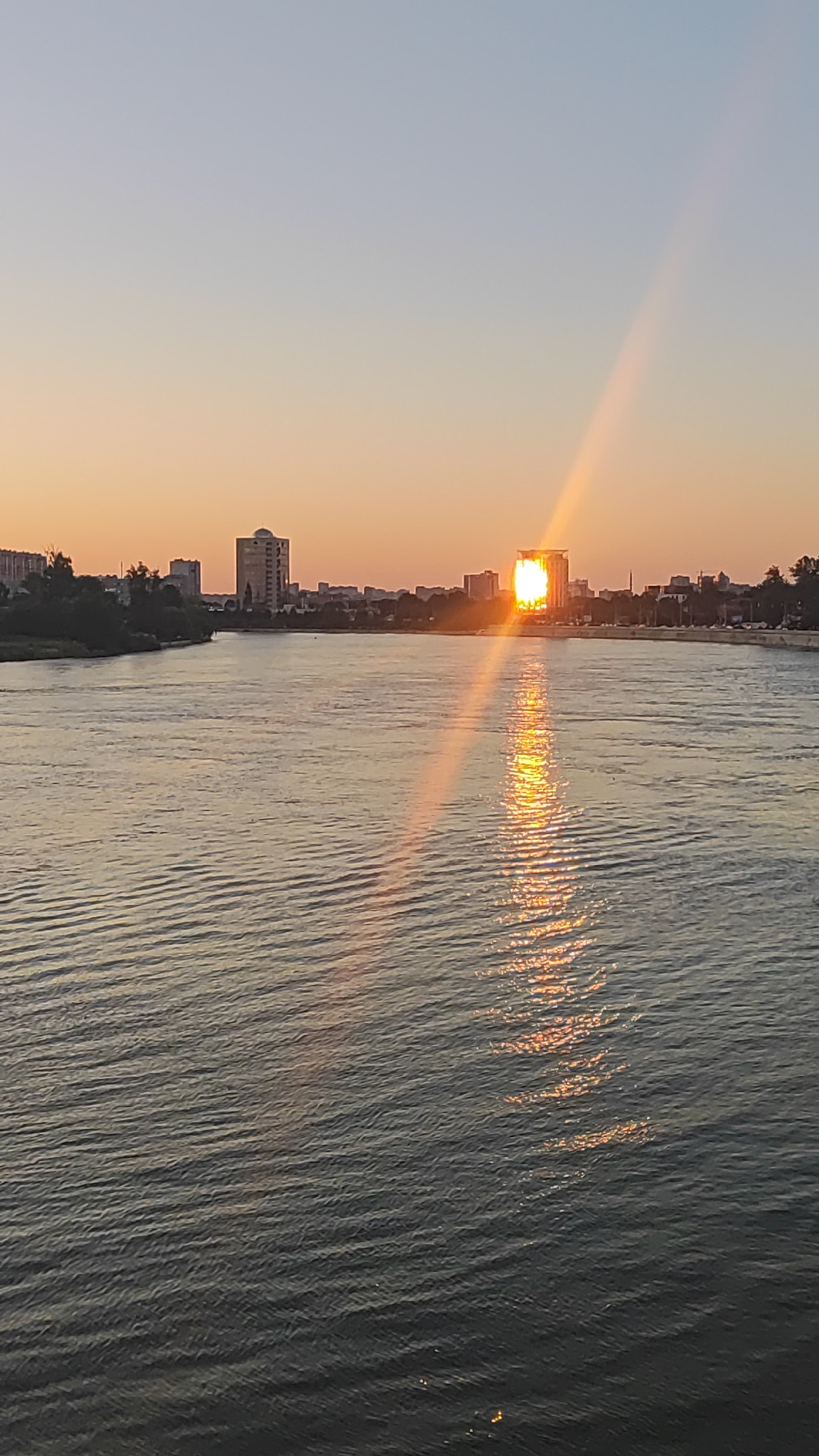 This is a vertical, medium-range, eye-level shot of a sunset over the Kuban River in Krasnodar, Russia. The sun, a bright orange orb, is setting behind a cluster of buildings on the far bank of the river, creating a strong glint on the water. The sky is a soft gradient of pale blue at the top to a warm orange near the horizon. The river occupies the majority of the foreground and midground, with gentle ripples reflecting the colors of the sunset. A line of trees and buildings can be seen along the far bank of the river, with the sun illuminating one prominent building with a large, rectangular, glass facade. The overall mood is serene and tranquil, with the natural beauty of the sunset dominating the scene. There are no people visible in the image.