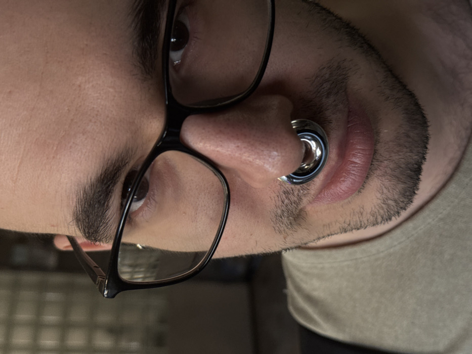 A close-up, eye-level portrait shows a young adult male looking directly at the camera with a subtle expression. He has dark hair, dark eyes, and a light complexion, with visible stubble around his chin and upper lip. He wears black-framed eyeglasses. A large, metallic, silver-toned circular septum piercing is prominently centered in his nose. He is wearing a light olive-green or beige crew-neck shirt. The background is shallowly focused and indistinct, appearing to be an indoor setting with muted tones and a hint of a rectangular grid pattern on the left. The specific location within Moscow, Russia, is not discernible from the image.