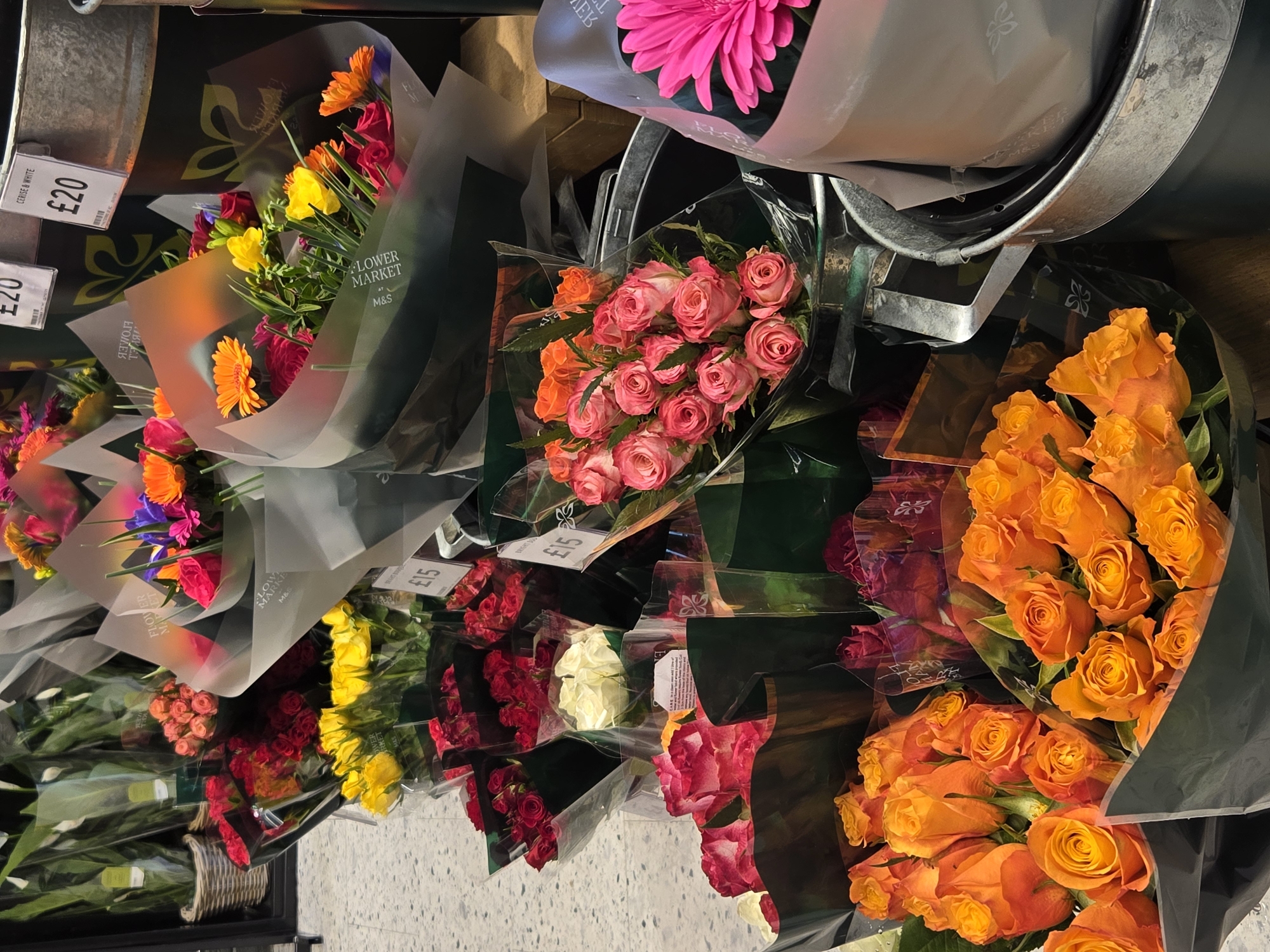 This image captures a vibrant, multi-tiered display of fresh flower bouquets inside a Marks & Spencer retail store, identifiable by "FLOWER MARKET M&S" branding visible on several wraps and the "£" currency symbol on price tags, placing the location within London, United Kingdom.
The foreground is dominated by numerous bouquets of orange roses, each wrapped in a dark green or black opaque film and a clear plastic sleeve. To the right, additional bouquets are displayed in galvanized metal buckets; these include light pink roses with white edges and a bright fuchsia pink gerbera daisy. Further back and slightly elevated, a variety of other bouquets are presented. These include prominent bunches of dark red roses, vibrant yellow roses, and white roses, all individually wrapped in similar clear plastic and opaque films. Mixed bouquets, containing flowers such as orange gerbera daisies, yellow freesias, and other colorful blooms in shades of red, pink, and purple, are also visible, some wrapped in translucent white paper. Price tags of "£20" and "£15" are visible on several bouquets, with one specifically stating "£20 CERISE & WHITE". The floor consists of light-colored, speckled square tiles. In the background, more plants, some in clear plastic sleeves and wicker baskets, are visible on shelves, confirming a retail environment. No individuals are present in the frame.