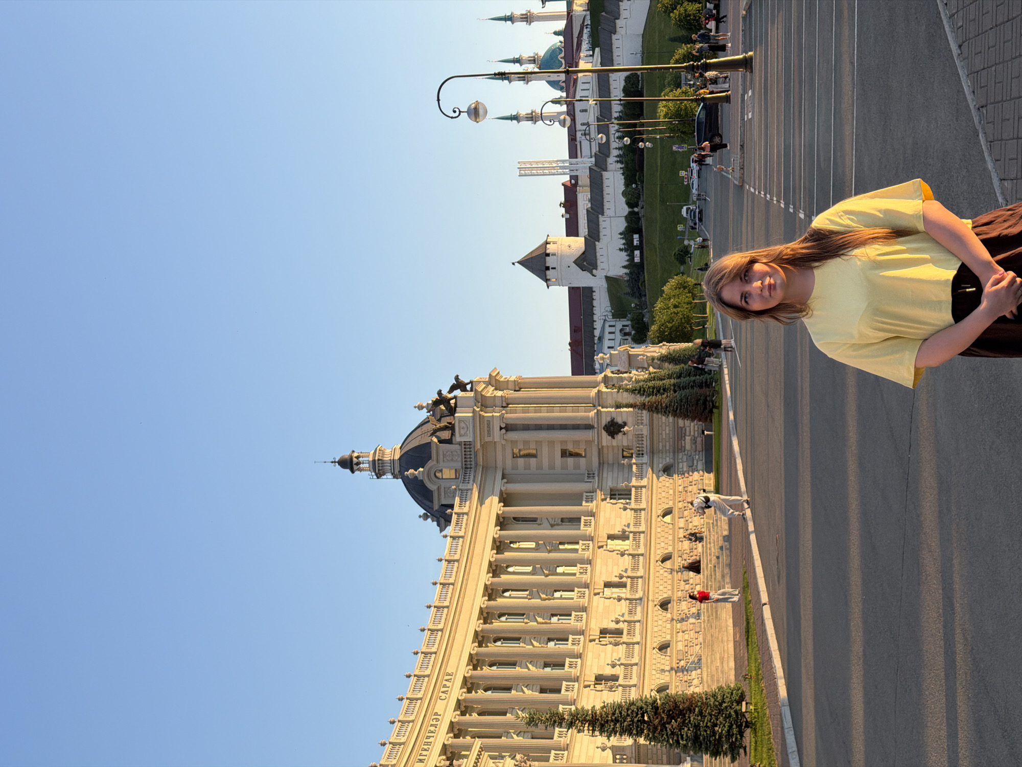 The image captures a street-level scene in Kazan, Russia, during the golden hour of a clear day. The foreground features a young adult female, approximately late teens to early twenties, with long, light brown hair, standing centrally and facing the camera. She wears a yellow short-sleeved t-shirt and a dark brown or black skirt, with her hands clasped in front. She stands on a paved area with a stone curb and visible road markings behind her.
To her left, dominating the midground, is the grand, light-colored neoclassical building identified as the Agricultural Palace (Ministry of Agriculture and Food of the Republic of Tatarstan). This structure features numerous tall columns, a large central dome topped with a finial, and architectural details including statues of eagles on its roofline. An inscription, partially visible as "ИГЕНЧЕЛЭР САРАЕ" (IGENCHELER SARAE, meaning "PALACE OF AGRICULTURALISTS" in Tatar), adorns the facade. Several evergreen trees are planted along the base of the building, and multiple individuals are visible on the steps and surrounding walkways, including one person in a red top and light pants, and another in white clothing.
Extending into the midground and background is a wide, paved street with traffic lanes and parking areas. Scattered vehicles and numerous pedestrians are present further down the street. Ornate streetlights with spherical lamps line the right side of the street. In the distant background, situated on a rise, are the white walls and towers of the Kazan Kremlin. Prominently visible within the Kremlin complex are the distinctive blue dome and multiple pointed minarets of the Kul Sharif Mosque, along with other white structures. The sky above is clear and light blue. Long, defined shadows are cast across the paved surfaces, indicating a low sun angle from the left side of the frame.