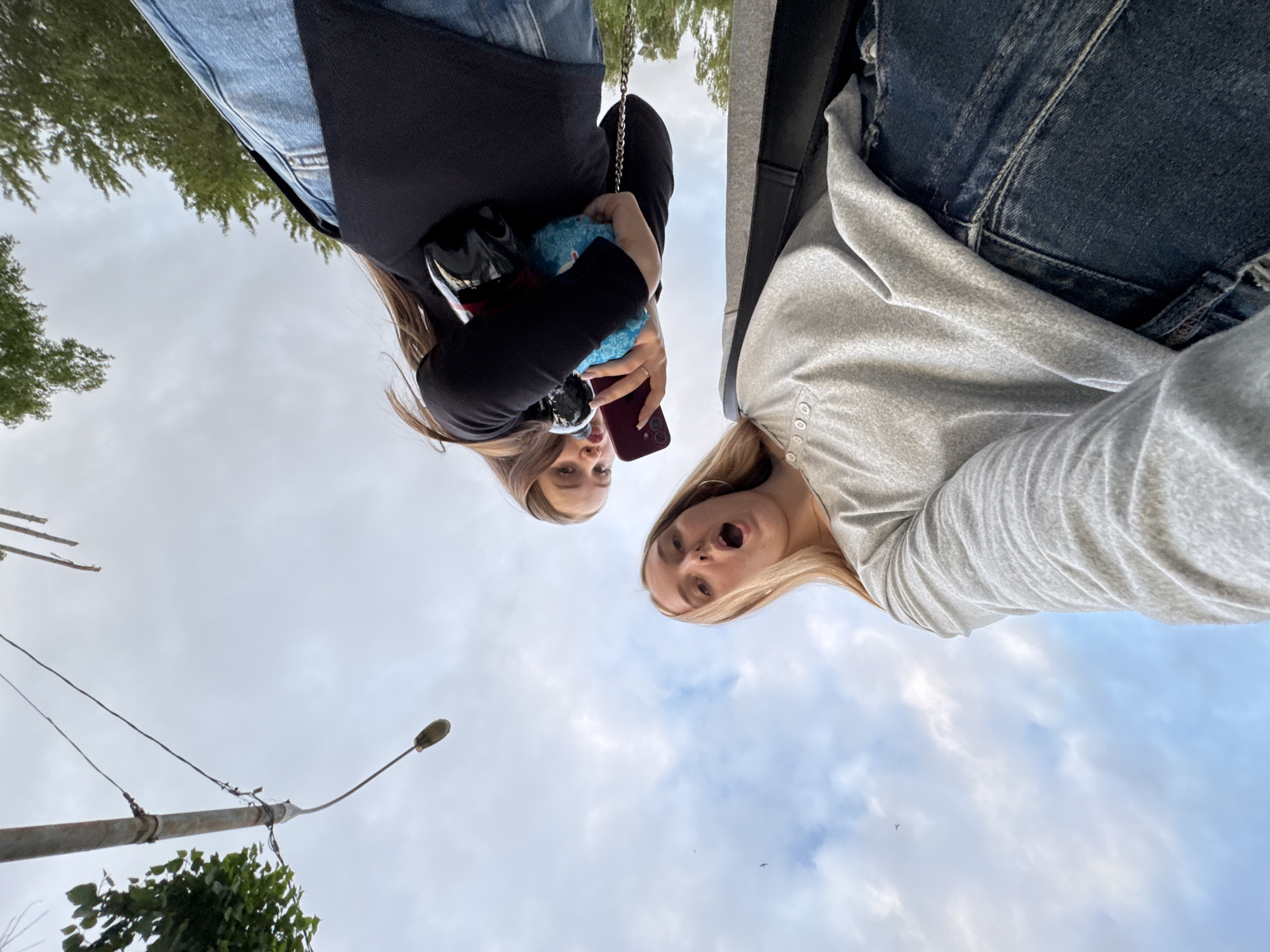 This image captures an upward-looking perspective of two young females in an outdoor urban or suburban setting within Perm, Russia, beneath a cloudy sky. The subject in the foreground, with long blonde hair, wears a light grey long-sleeved shirt featuring button placket details and dark blue denim jeans; her mouth is open, conveying an expression of surprise or speech. Positioned behind and above her is a second female with long brown hair, dressed in a black long-sleeved top and dark blue denim jeans. She holds a red smartphone in her left hand and a blue, possibly plush, item or small bag with a chain strap in her right hand. Both individuals are looking downwards towards the camera, suggesting a low-angle selfie or a photograph taken from a prone position. The upper-left background shows a grey utility pole with wires, from which a dark, elongated object dangles. Green foliage from trees or bushes is visible on the left near the pole and also in the upper right. The sky is predominantly light grey with scattered white clouds, and a small bird is visible in flight in the mid-left portion of the sky.