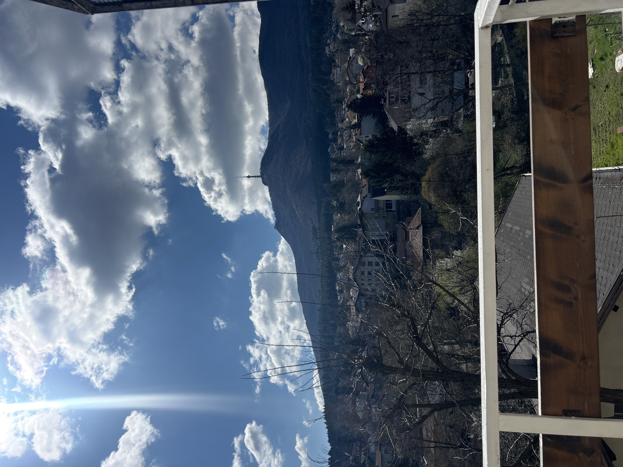 The image depicts a landscape view from a high vantage point, possibly a balcony. The sky is predominantly blue with scattered, puffy white clouds, indicating a sunny day. A mountain dominates the midground, its slopes covered with a dense forest that transitions into a town or settlement. Buildings, mostly residential, are clustered along the base of the mountain and extend into the valley. In the foreground, a section of a balcony railing and support structure is visible. The railing is white, and the support is made of stained wood. The environment is serene, suggesting a residential area with natural surroundings.
The location appears to be in or near the city of Sofia, Bulgaria, given the architecture and mountainous backdrop.