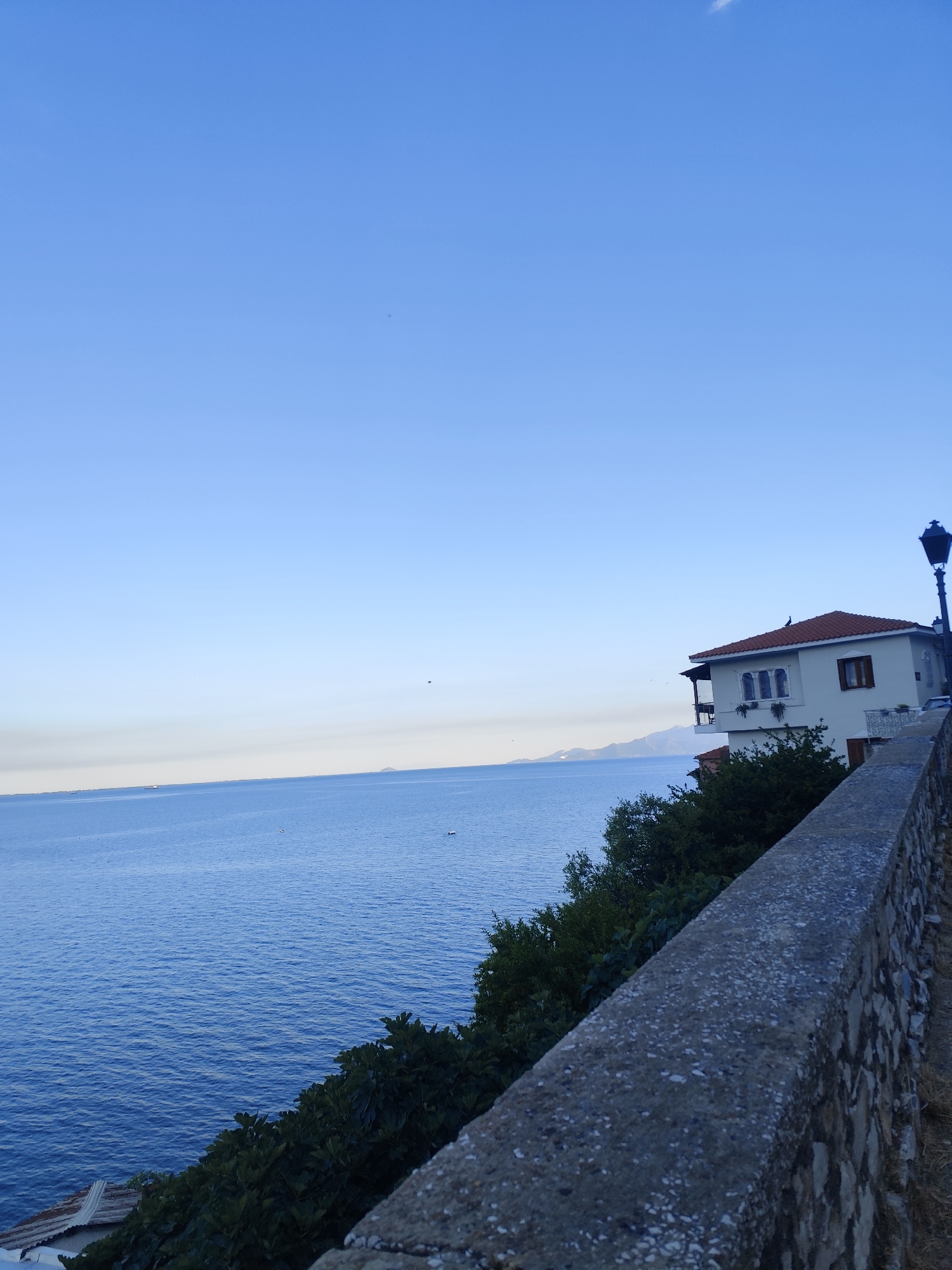 A coastal view in Kavala, Greece, features a tranquil blue sea meeting a clear, pale blue sky. The horizon line is sharp, with distant landmasses visible on the left. A white building with a red-tiled roof, adorned with arched windows and balconies, sits atop a lush green slope. A vintage-style black lamppost stands to the right of the building. In the foreground, a textured stone wall runs diagonally across the scene, separating the viewer from the vibrant vegetation below. The overall impression is one of serene beauty and calm, possibly during early morning or late afternoon given the soft lighting.