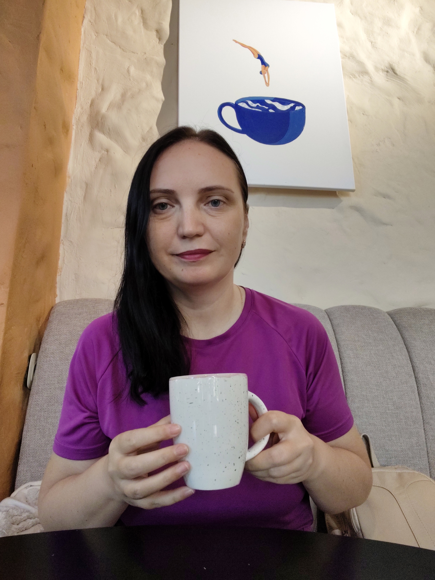 A medium shot captures a woman with long, dark hair and a purple t-shirt sitting on a light grey upholstered couch. She is holding a white speckled mug with both hands. In the background, a painting of a diver jumping into a blue coffee cup hangs on a textured, off-white wall. To the left of the painting, another wall is visible, painted in a warm, sandy tone with a rough texture. The overall setting appears to be a cafe or lounge, possibly in Sochi, Russia, given the decorative wall textures often found in establishments there. The woman is looking directly at the camera with a neutral expression. The lighting is soft and diffused, suggesting an indoor environment.