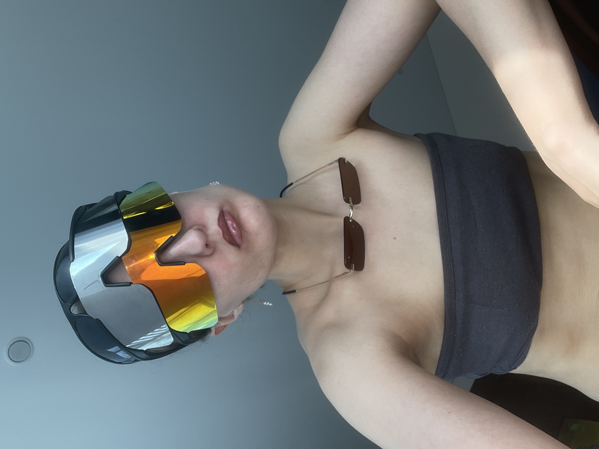 A woman is positioned with her body angled towards the camera, her head tilted back. She wears futuristic-looking glasses with silver and orange lenses. Another pair of sunglasses rests on her chest, suspended by a chain or cord. She is wearing a dark gray strapless top. The background is a plain, light gray wall. There is no visible evidence that the image was taken in Подшивалово, Russia.