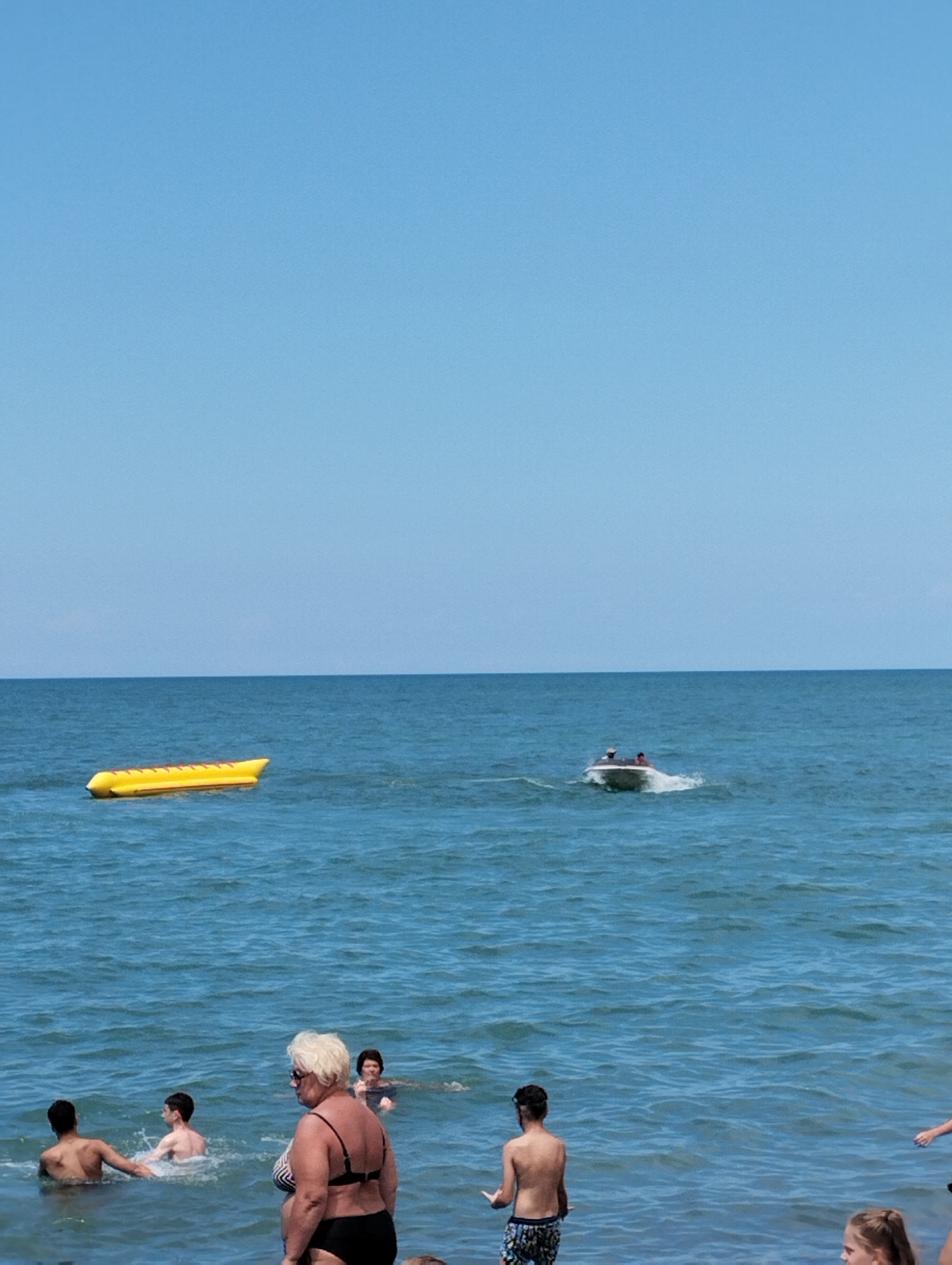 In a bright, clear sky over a calm, blue sea, people enjoy a summer day at the beach. A yellow inflatable banana boat is visible in the distance, resting on the water. Closer to shore, a small motorboat with two occupants navigates the waves. Several people are in the water near the foreground. A woman with blonde hair, wearing a striped bikini, stands facing away from the camera, looking out at the sea. Behind her, two boys are wading in the water. Further out, another person is swimming. The scene suggests a relaxed atmosphere on a sunny day. The clear blue sky indicates good weather, and the presence of people in swimwear and water activities confirms it is likely daytime. No visible text is present in the image. The location is Batumi, Georgia.