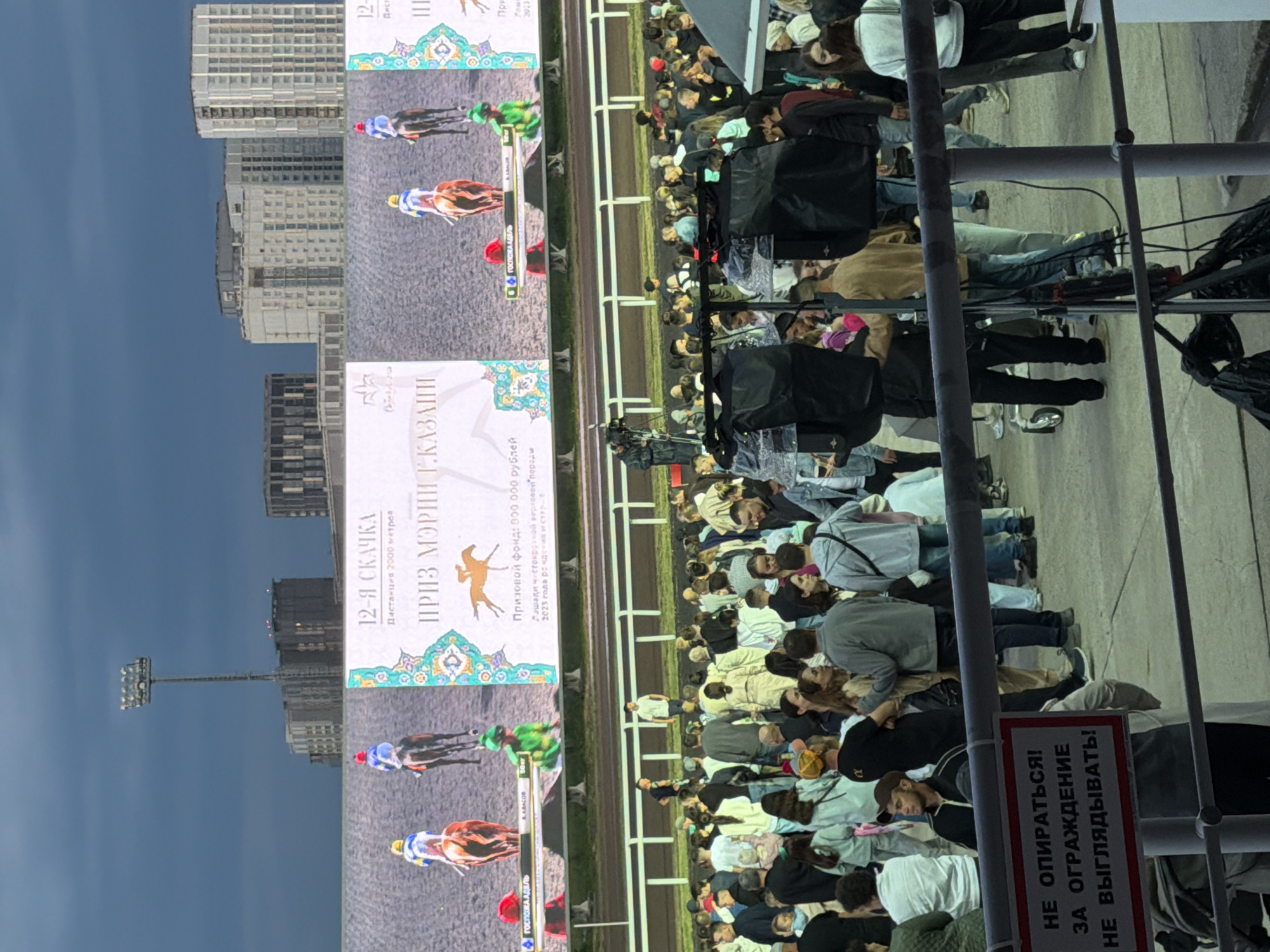The image captures a horse race event, likely at a racetrack, under an overcast sky. A large digital screen displays a live feed of horses and jockeys racing, with text in Russian indicating it's the "12th Race" and the "Priz of the President of Tatarstan." The screen also shows a prize fund of 500,000 rubles. In the foreground, a crowd of spectators is gathered, facing the screen and the track. Some individuals are holding phones, possibly recording or taking pictures. A few people are standing near a safety barrier. The environment appears to be outdoors, with a paved area for the spectators and a fenced track. Several floodlights are visible in the background, suggesting the event might be taking place in the evening or under low light conditions. The overall atmosphere seems to be one of excitement and engagement with the horse race.