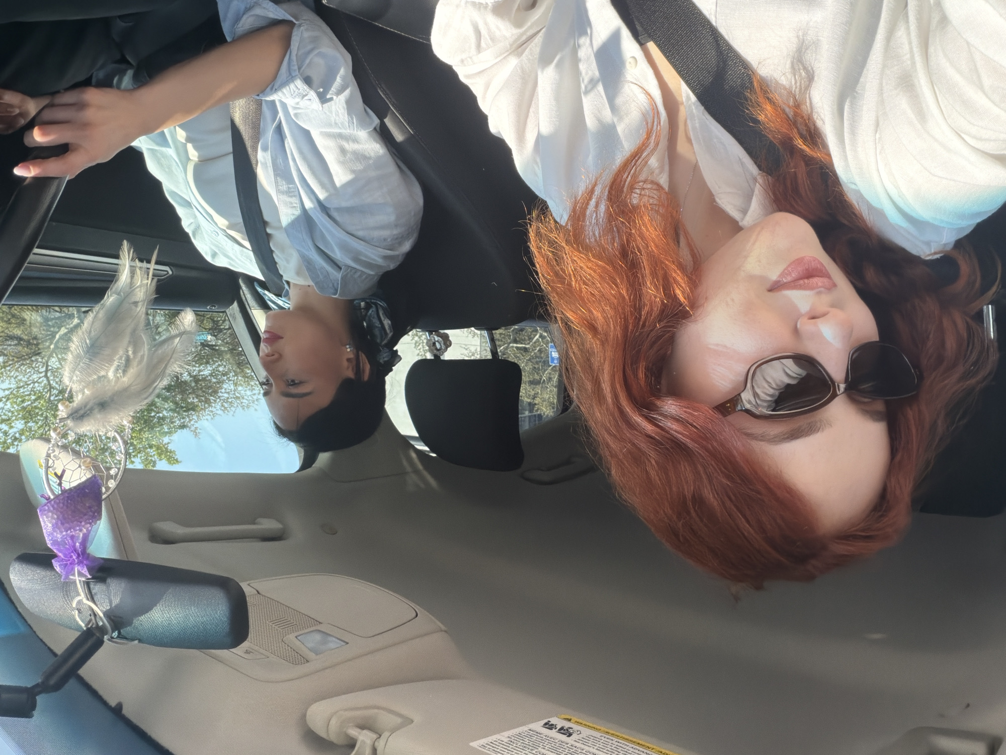The image captures two women inside a car on a bright, sunny day in Tbilisi, Georgia. The perspective is from the rear of the vehicle, looking forward.
In the front passenger seat, a woman with vibrant reddish-brown hair is visible. She wears dark sunglasses, which reflect the bright sky, and a light-colored, possibly white, collared shirt, secured by a seatbelt. Her face is slightly turned upwards with a neutral expression, and sunlight strongly illuminates her features and hair.
The driver, a woman with dark hair, is seen in profile. She wears a light blue button-up shirt over a lighter top, also with a seatbelt across her chest. Her left hand rests on the dashboard or steering wheel. Her gaze appears directed forward.
Hanging from the rearview mirror is a decorative ornament resembling a dreamcatcher, featuring white feathers and distinct purple mesh or fabric accents. The car's interior, including the beige headliner, sun visors, and parts of the dashboard, is clearly visible. Through the car windows, a clear blue sky and the tops of green trees are seen, indicating an outdoor, natural setting.
The strong sunlight creates noticeable highlights and shadows throughout the image, emphasizing the sunny weather. On the passenger-side sun visor, a white warning label with small black text is partially legible, including fragments such as "WARNING" and phrases related to "AIRBAGS." The scene depicts a calm moment, likely during travel or a stop, within the vehicle.