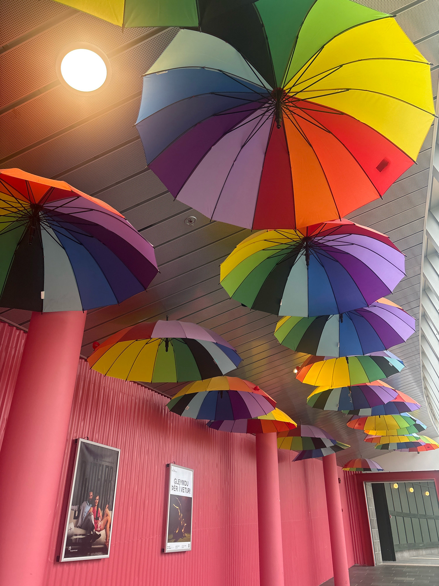The image shows a brightly colored ceiling installation of numerous rainbow-colored umbrellas suspended at varying heights, creating a vibrant canopy. The umbrellas are made of fabric and have black handles. They are arranged in a decorative manner above a pink corrugated wall. On the wall, there are two framed posters. The poster on the left depicts two individuals seated, with text below. The poster on the right has text at the top and an image of a dancer or performer below. The environment appears to be indoors, possibly a public space or a venue, with a ceiling featuring a ventilation grate and a circular light fixture. The overall impression is festive and artistic. This location is the Nordic House in Reykjavik, Iceland.