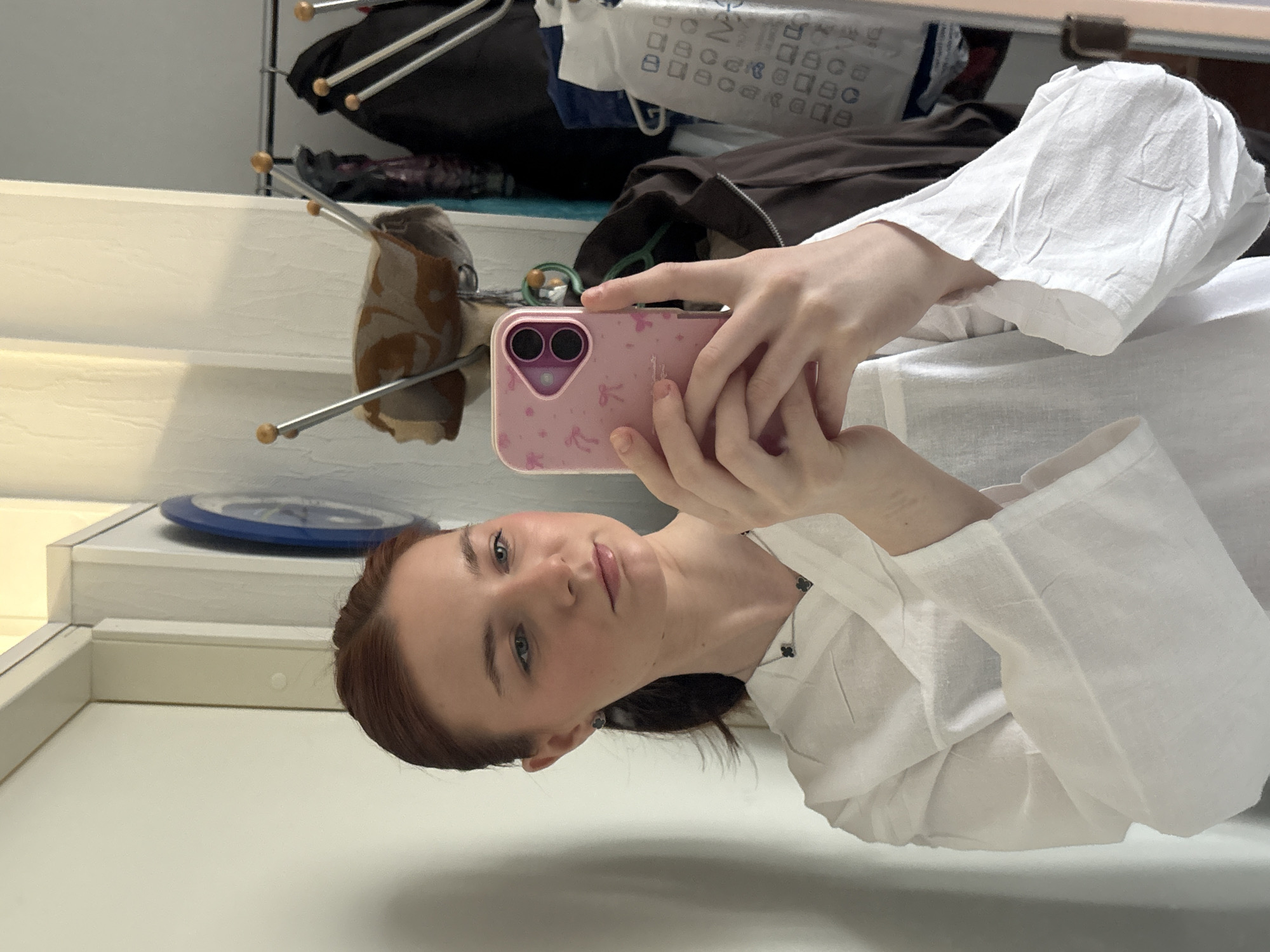 A young woman with red hair and blue eyes is taking a selfie with a pink smartphone. She is wearing a white long-sleeved shirt and a black necklace with clover-shaped pendants. In the background, there is a white shelf with a blue plate and a clothing rack with various items of clothing, including a brown jacket, a black bag, and a white bag with Cyrillic text. The location appears to be indoors, possibly in a home. There is no indication of Saint Petersburg, Russia in the image.