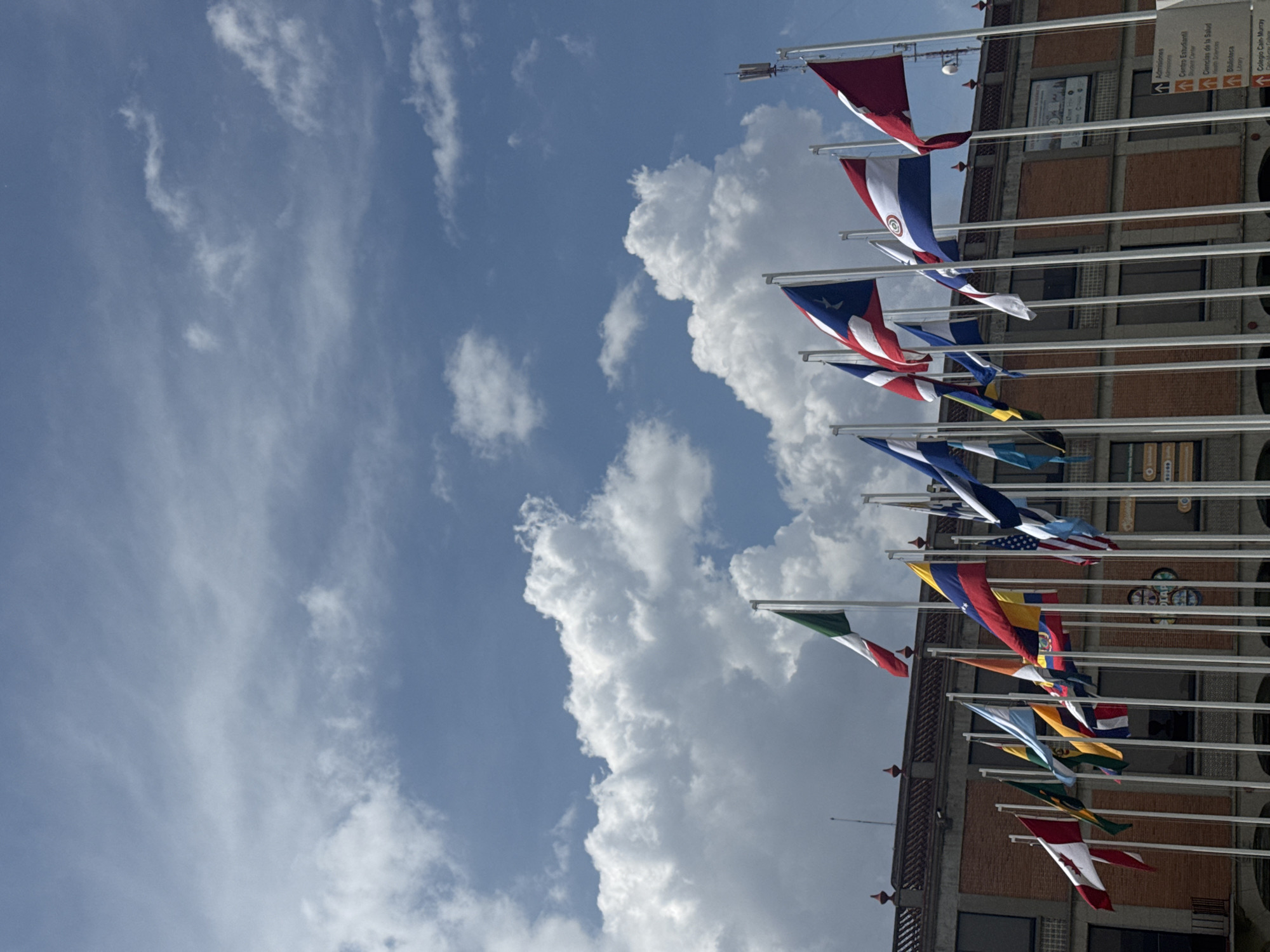 The image shows a view of the sky with white clouds and a building facade with flagpoles displaying various national flags. The building is a brick structure with concrete columns. There is a sign in Spanish that says "Admisiones, Centro Estudiant, Ciencias de la Salud, Biblioteca." The sign is likely referring to the administrative offices of the building. The flags include the flags of Puerto Rico, Paraguay, the United States, and other nations. The location is in San Andrés Cholula, Mexico.