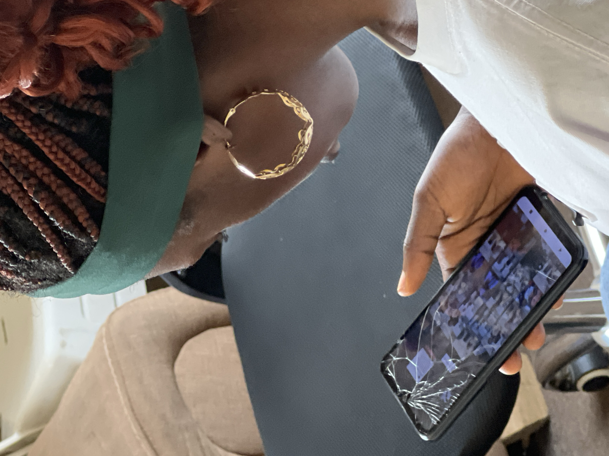 An over-the-shoulder medium shot shows a Black woman sitting on a black chair, looking down at a cracked smartphone in her hands. She is wearing a green headband, large gold hoop earrings, and a white t-shirt. Her braided hair is visible at the top of the frame, with some curly reddish-brown hair framing her face. The smartphone screen displays a mosaic of images, the details of which are obscured by cracks across the screen. The background includes a beige armchair and a dark, textured surface, possibly a table or desk. The lighting is natural and appears to be coming from the right. There is no visible indication of the city of Jos, Nigeria.