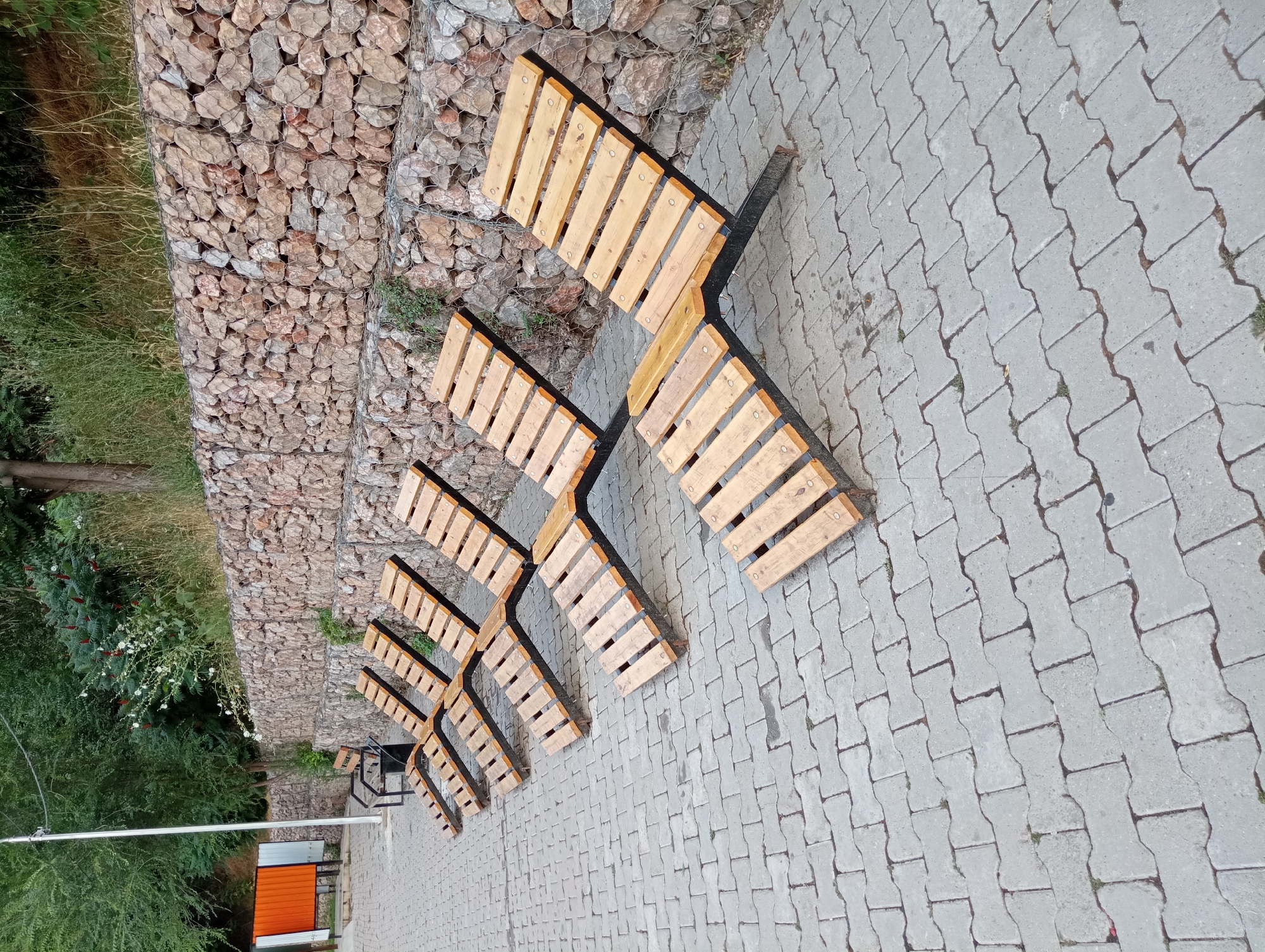 The image, taken in Shymkent, Kazakhstan, captures an outdoor scene featuring a pathway paved with interlocking concrete blocks. Alongside the pathway, a series of modern wooden benches with dark metal frames are arranged in a staggered pattern. The benches are positioned adjacent to a sloped embankment constructed with gabion baskets filled with rocks, covered in wire mesh. Lush green vegetation, including grass and trees, grows along the base of the embankment and in the background. A metal pole and a structure with an orange door are partially visible in the lower left portion of the frame. The lighting suggests it is daytime. There are no people present in the image.