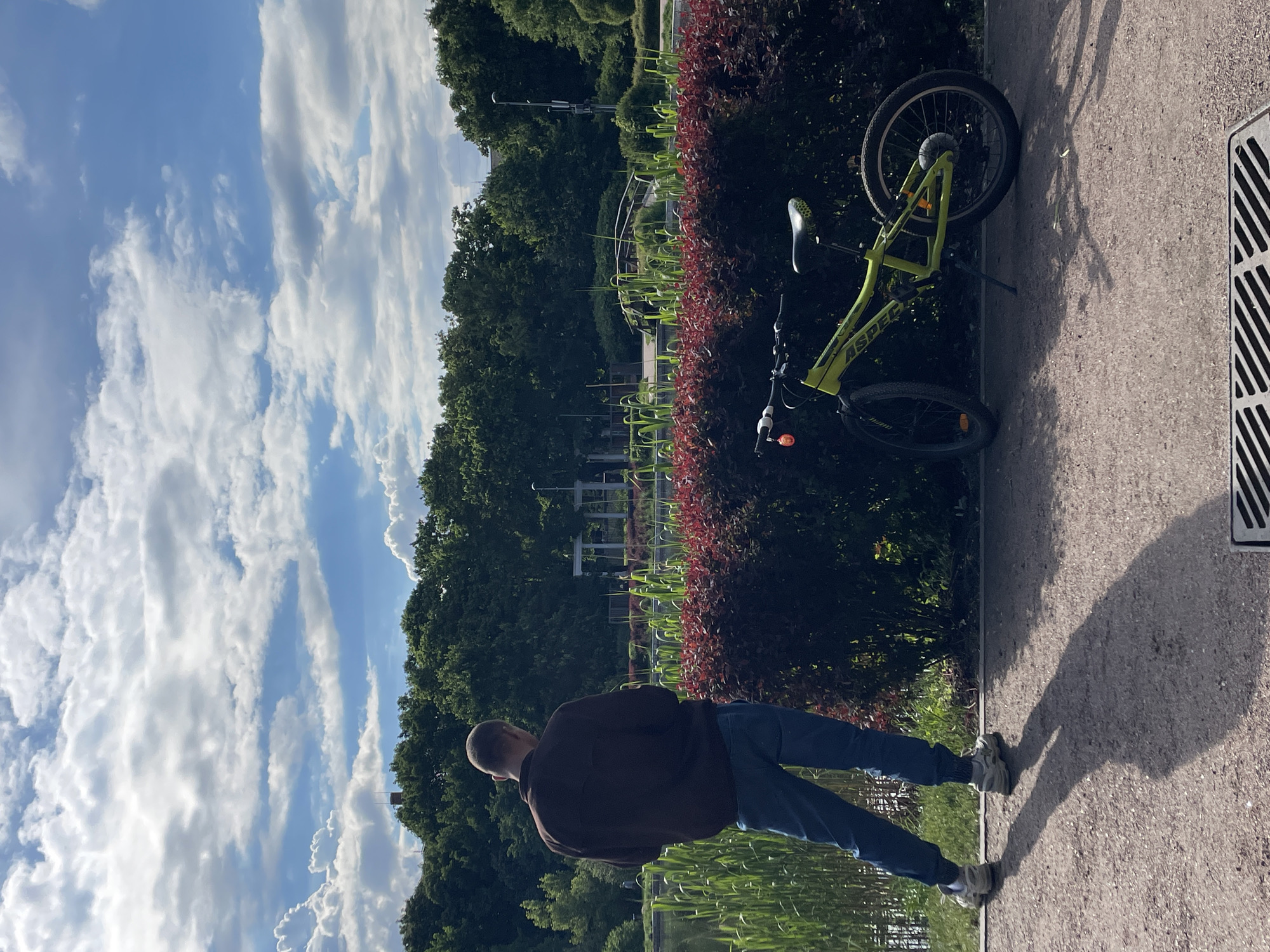A man in a brown shirt and blue jeans is standing near a body of water, possibly a lake or pond. In the background, there is a lush green park with a variety of trees and shrubs. A green bicycle is leaning against a hedge in the middle-ground. The sky is partly cloudy with white clouds and patches of blue. The ground is paved. Based on the presence of specific structures, vegetation, and water features similar to those in the image, the location is possibly Khimki, Russia.