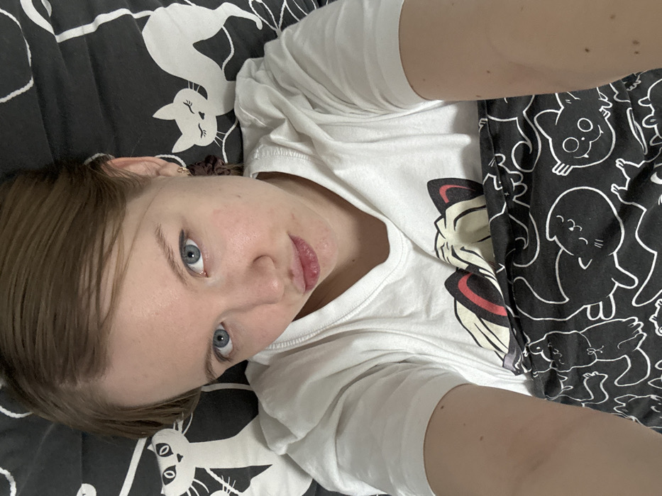 A person with short brown hair and blue eyes lies on a bed. The bedspread features a pattern of white cats on a black background. The person wears a white t-shirt with a cartoon cat graphic. The person is also wearing black pants with cartoon cat graphics. The photo is taken from a slightly high angle, with the person looking up at the camera.