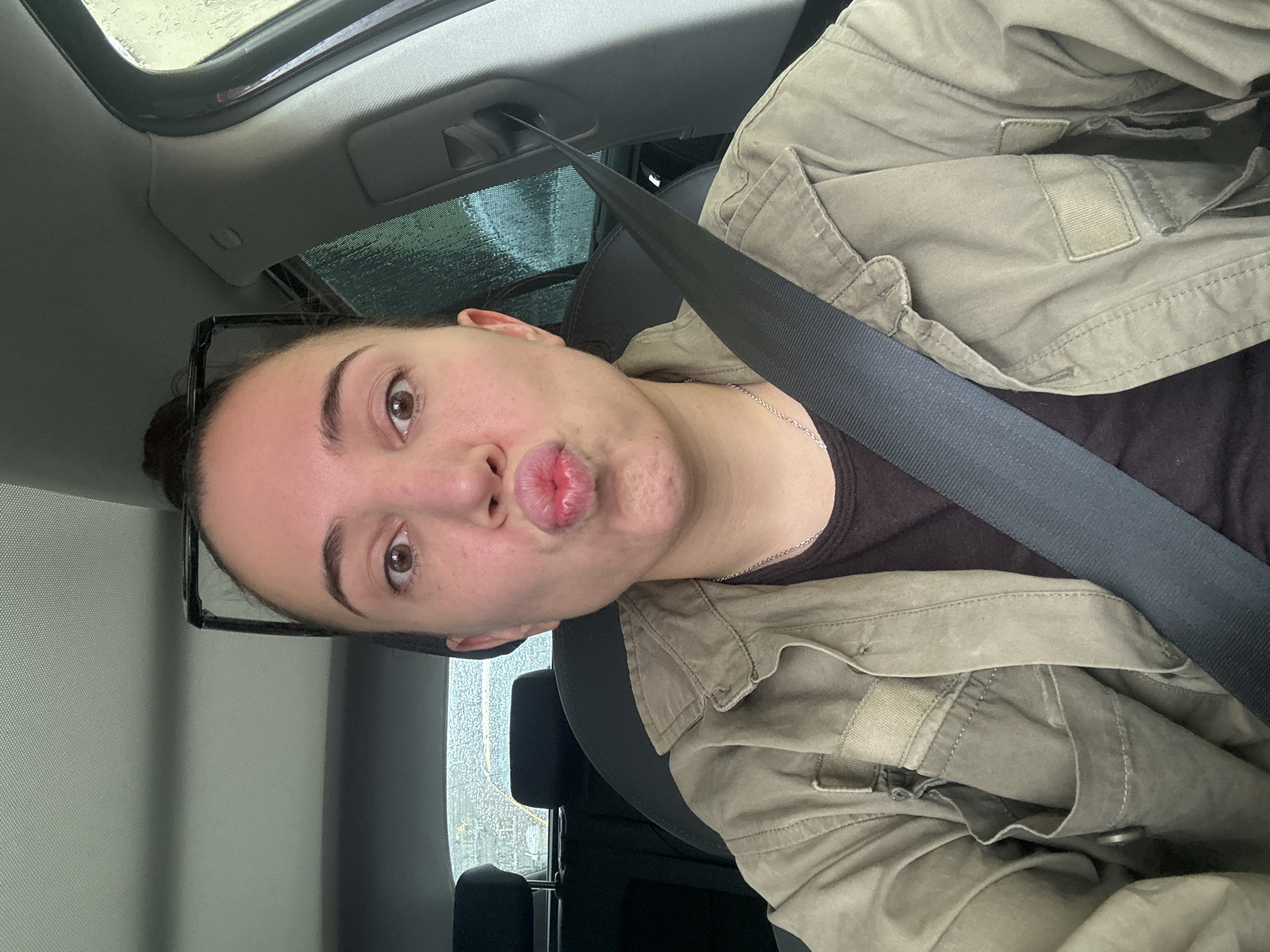 A woman with her hair in a bun, wearing a brown shirt and a green jacket with a seatbelt across her chest, is in the driver's seat of a car. She is puckering her lips and looking at the camera. The background shows the interior of the car, including the dashboard and a rainy window. The scene is indoors, inside a vehicle, and the exterior environment appears to be rainy. There is no information in the image to suggest a location in Moscow, Russia.