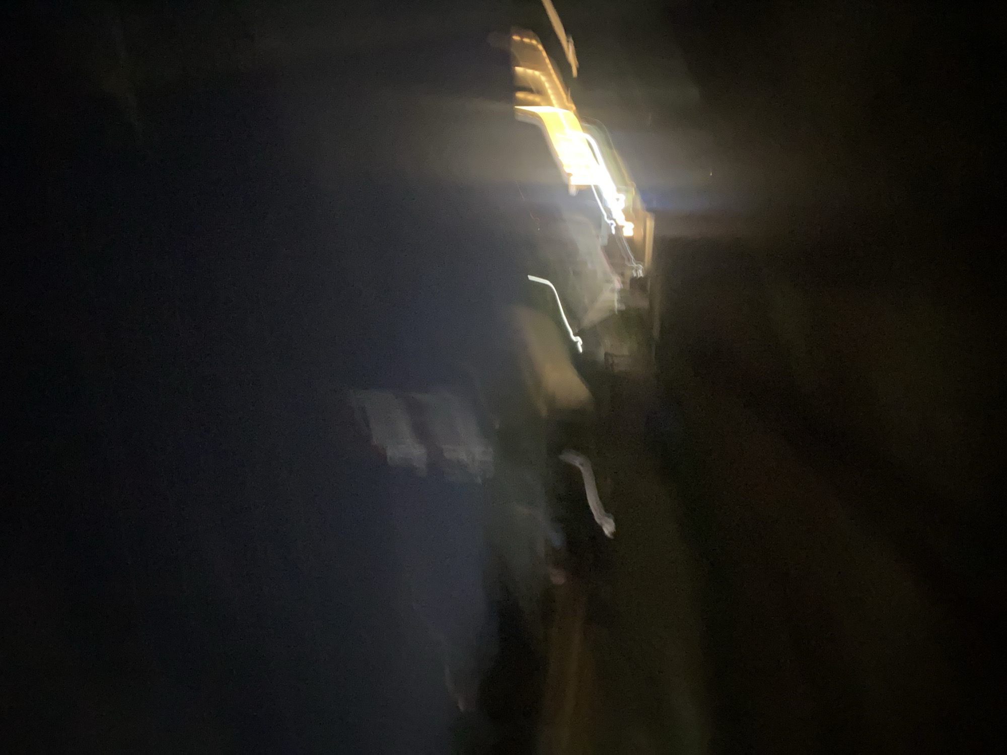 The image depicts a blurry, abstract scene at night, suggesting motion or a low-light photograph. Several streaks of light are visible, predominantly yellow and white, possibly from streetlights, vehicle headlights, or illuminated buildings. These lights are elongated and fragmented due to movement or a long exposure. In the background, there are indistinct shapes that could represent structures, though their details are obscured. The overall impression is one of a dynamic, possibly urban, environment at night. Without more specific visual cues or context, pinpointing the exact location within Jos, Nigeria is not possible.