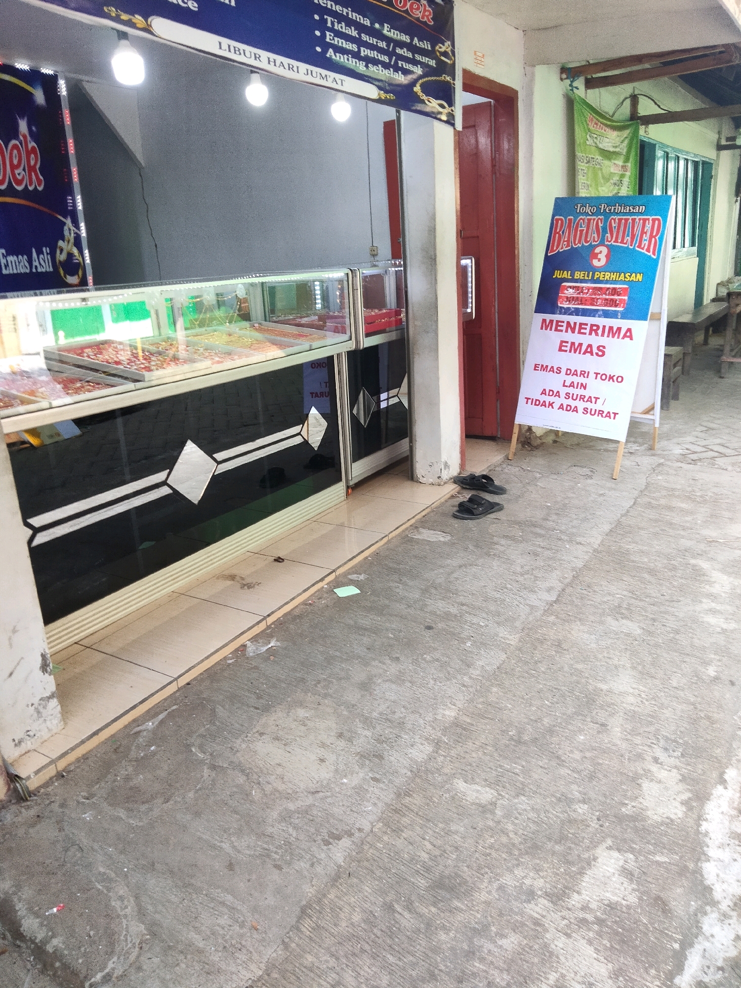 The image depicts the exterior of a jewelry store located in Sawahan, Indonesia. The store appears to be a small, local business with a display case showcasing various jewelry items. A sign in front of the store advertises "Bagus Silver" and states that they buy gold, even if it's from other stores or without a receipt. The store is situated on a street with a concrete sidewalk. There are a pair of black sandals placed on the ground outside the store, and a small green piece of paper is visible on the sidewalk. The overall environment suggests a modest commercial area.