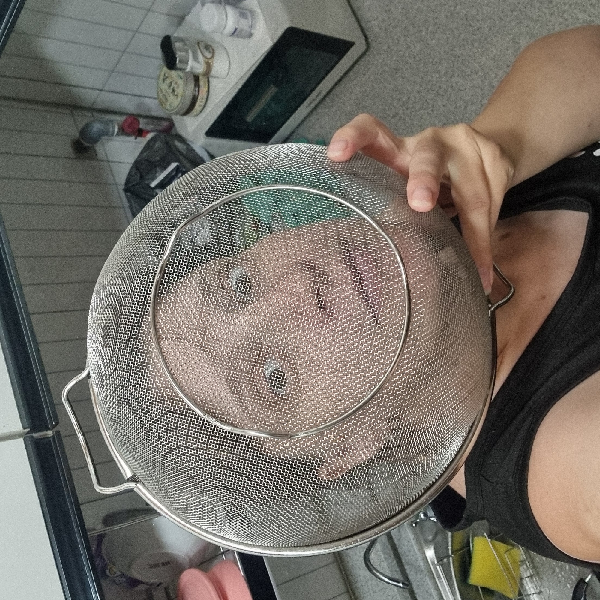 The image depicts a person's face seen through a metal sieve in a kitchen setting. The person is wearing a black tank top, and their face is distorted by the mesh of the sieve, with visible eyes, nose, and mouth. The background shows kitchen counters, a microwave, jars, and a sink with a sponge. The location is identified as Dunnae-myeon, South Korea.