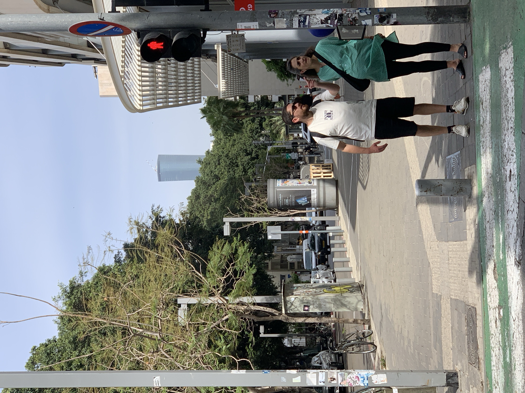 The image captures a street scene in Tel Aviv, Israel. Two pedestrians, a man and a woman, stand on a sidewalk near a pedestrian crossing marked with green stripes. The man wears a white t-shirt, black shorts, sunglasses, and white sneakers. The woman is dressed in a green top, black leggings, and black sandals. A red "wait" signal is illuminated at a pedestrian traffic light overhead. In the background, lush green trees line the street, and a tall skyscraper is visible against a clear blue sky. Bicycles are parked near a tree, and there are parked cars on the road behind the sidewalk. Street signs and advertisements are affixed to lampposts and structures along the sidewalk.