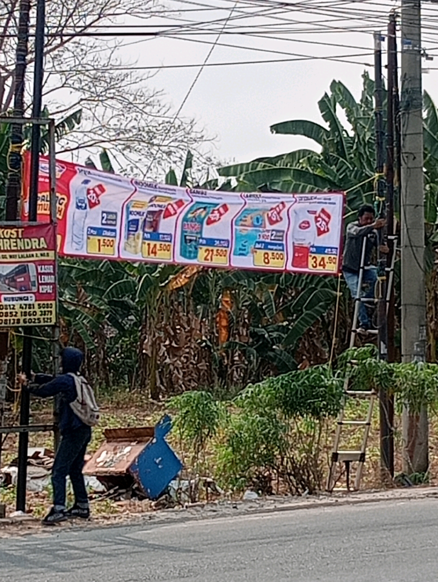 In Bandar Lampung, Indonesia, two men are working on a street-level banner advertising grocery products. One man, wearing a dark jacket and jeans, is on a stepladder attached to a utility pole, adjusting the banner. The other man, wearing a dark hoodie and backpack, stands on the ground, holding onto a different utility pole and appearing to be assisting with the banner's placement. The banner displays various products, including bottled water and milk, with prices such as "1.500", "8.500", and "34.90". Behind the banner, lush green banana trees are visible. To the left, a smaller red and white sign with text including "KOST HENDRA" and contact numbers is attached to a pole. The scene is set along a paved road, with debris and foliage present on the roadside.
