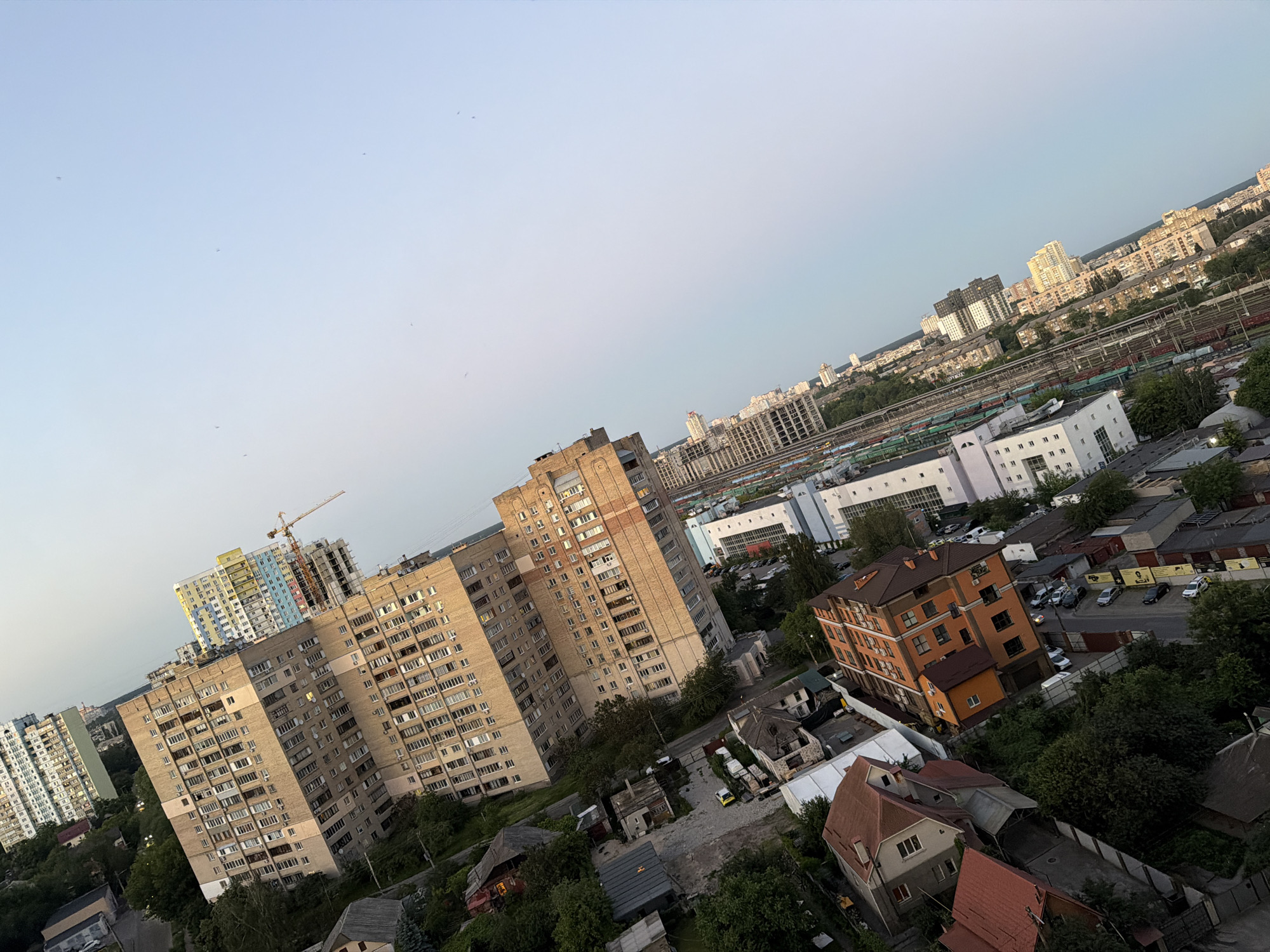 The image shows a panoramic view of a city skyline with numerous residential buildings, some under construction, and infrastructure like railway tracks. The perspective suggests an elevated viewpoint, possibly from a high-rise building or drone. The buildings are diverse in height and architectural style, ranging from older, multi-story apartment blocks to newer, modern constructions. A prominent feature is a construction crane atop a building that appears to be under development. The presence of railway tracks with several train cars indicates a transportation hub or industrial area within the city. The scene is illuminated by natural light, suggesting it was taken during the day, likely in the late afternoon or early evening due to the soft lighting. The sky is clear with a pale blue hue, and there are a few specks that could be birds or distant aircraft. The environment appears to be a densely populated urban area with greenery interspersed among the buildings, suggesting parks or treed areas within the city. The overall impression is one of a sprawling metropolitan landscape with ongoing development and activity. Based on the distinctive architecture of the buildings and the urban planning visible, this scene is likely located in Kyiv, Ukraine, possibly in a district characterized by a mix of older and newer residential developments and proximity to transportation infrastructure.