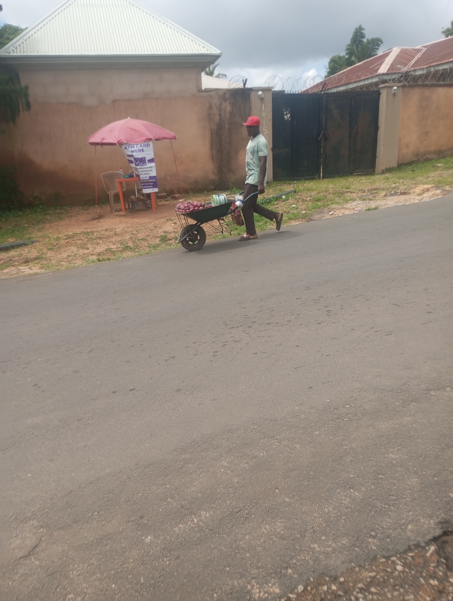 A man wearing a light green t-shirt and brown pants, with a red cap, is pushing a wheelbarrow full of red onions down a paved road in Jos, Nigeria. The wheelbarrow is partially obscured by the man, but several onions are visible in its basin. To the left of the road, there is a small market stall with a pink umbrella, an orange table, and a chair. A sign on the table advertises ATM card services. Behind the market stall, there is a beige building with a corrugated metal roof. To the right of the road, there is a wall with barbed wire on top and a black metal gate. The sky is overcast.