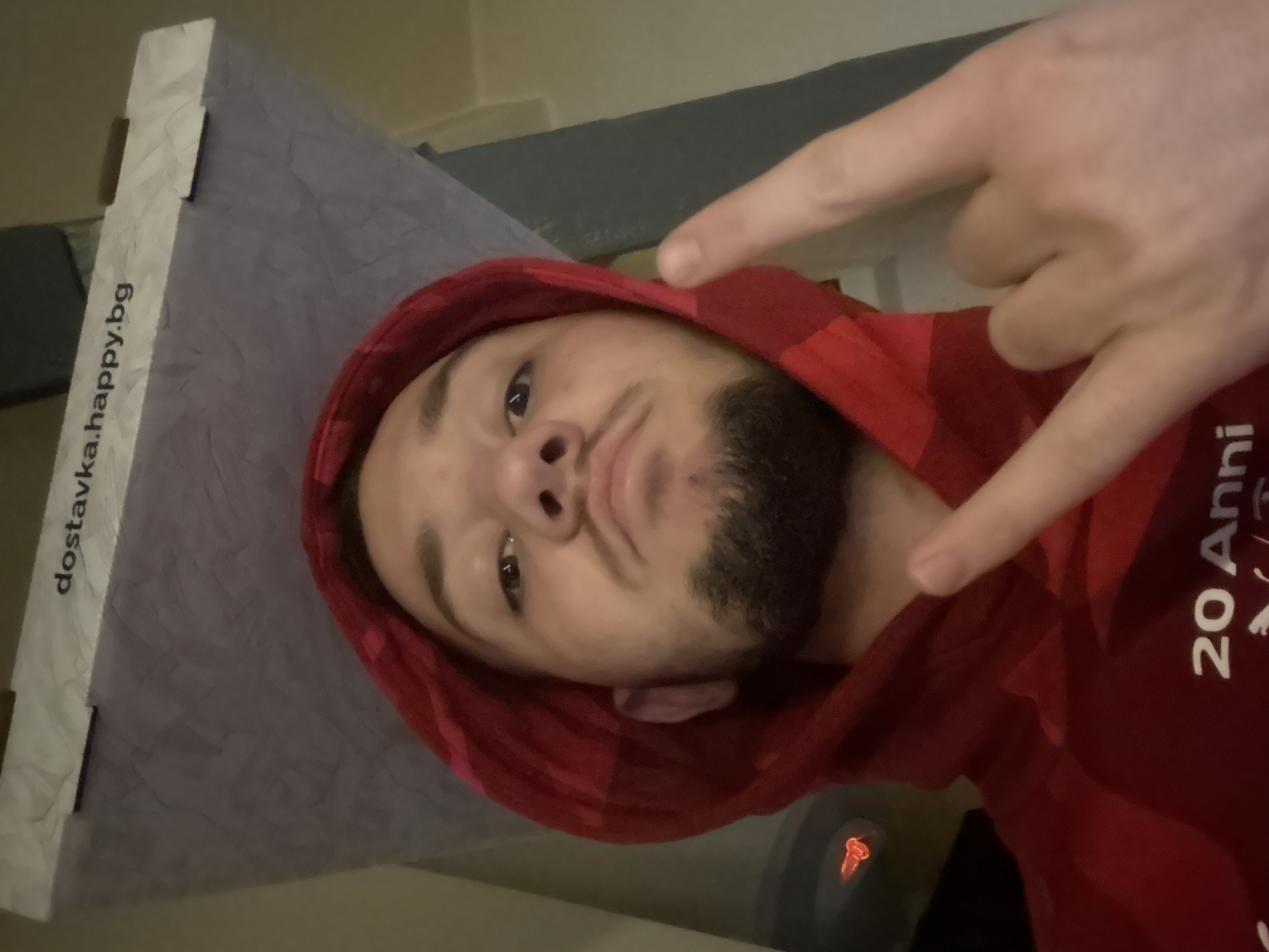 A man with a beard and a red hooded sweatshirt is making a "rock on" hand gesture. He is lying down, with his head resting on a gray textured surface with the text "dostavka.happy.bg" visible on the side. To the right of the man, the text "20 Anni" can be seen on the sweatshirt. The background includes a gray surface, possibly a bed or couch, and a small red light, possibly from a gaming headset. While the text on the gray surface indicates the delivery service "happy.bg," which is a company in Bulgaria, the location within Sofia is not directly discernible from the image.