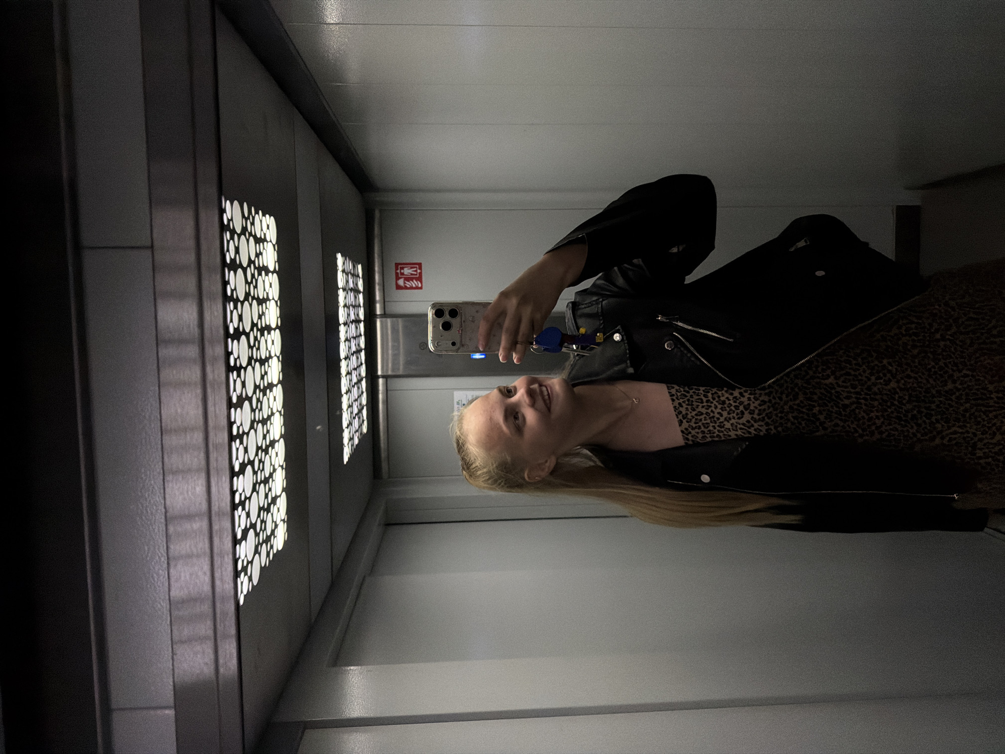 A woman is taking a selfie in an elevator. She is wearing a black leather jacket over a leopard print dress. She is holding a mobile phone in her right hand and looking at the screen. The elevator has decorative lighting panels on one wall. The other wall has a fire safety sign. The location appears to be inside a building, but nothing in the image indicates it is in Izhevsk, Russia.