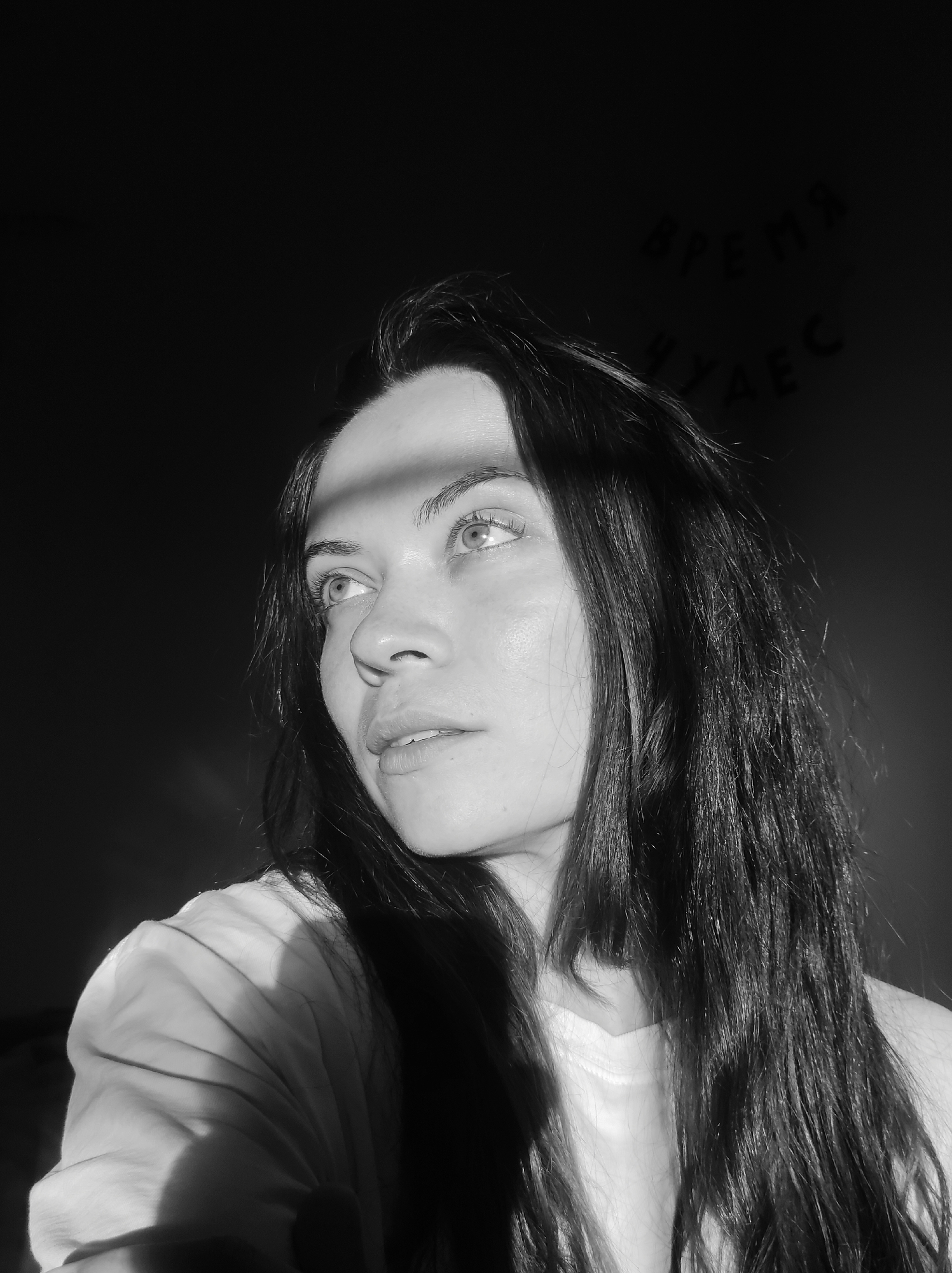 A high-contrast, black and white photograph depicts a young adult female captured from the chest up, positioned off-center to the right, in the city of Unknown, Unknown. She has long, dark, disheveled hair that partially obscures her right shoulder and falls over her left. Her head is slightly tilted, and she gazes upwards and to the viewer's left with a pensive expression; her lips are slightly parted. A strong, directional light source illuminates her from the upper left, casting a distinct shadow band horizontally across her left eye and forehead, and creating pronounced highlights and shadows on her facial features and the folds of her light-colored shirt on her right shoulder. The background is largely dark and indistinct. In the upper right quadrant, faint, lighter-toned Cyrillic text is visible against the dark background, reading "ВРЕМЯ ЧУДЕС," which translates to "TIME OF MIRACLES."