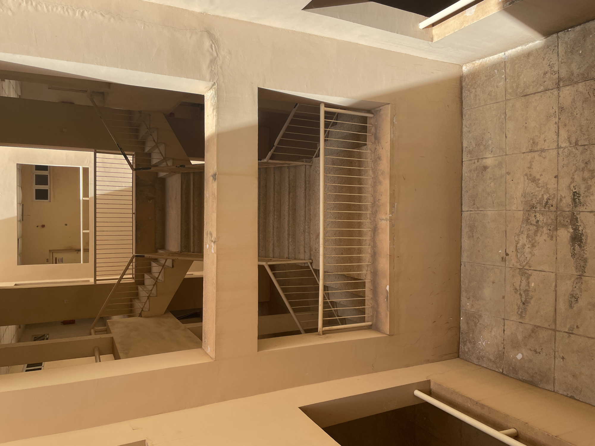This image depicts the interior courtyard of a multi-story building, likely an apartment complex or administrative building. The architecture features prominent beige concrete structures with open-air walkways and staircases. Two sets of outdoor staircases are visible, one on the upper level and another on a middle level, both equipped with simple metal railings. The staircases appear to be made of concrete. The courtyard floor is tiled with large, square, light brown tiles, some of which show signs of wear and staining. The lighting suggests it is daytime, with sunlight casting distinct shadows on the walls and floors, indicating an open-air environment. There are no people visible in the image. The overall impression is one of a functional, somewhat austere, urban building. The prompt mentions Kabala, Nigeria, but there is no visual information within the image to confirm or deny this location.