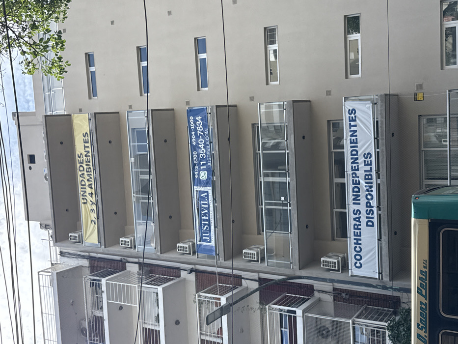 The image captures a slightly upward-angled view of the facade of a multi-story building, likely an apartment or commercial complex, under bright daylight. The building's exterior is a light, neutral color, possibly beige or off-white, and features multiple vertical windows that are tall and narrow. Below many of these windows, there are individual balconies or glass-enclosed extensions. Several white, rectangular outdoor air conditioning units are visibly mounted on the ledges or below these balconies.
Three large, vertical banners are prominently displayed on the building's facade, advertising property units and parking spaces.
1. The leftmost banner is yellow with black text, reading: "UNIDADES" (Units) and "2, 3 y 4 AMBIENTES" (2, 3 and 4 rooms/environments).
2. The central banner is blue with white text and a white logo, featuring contact information for a real estate entity. It displays: "JUSTEVILA", two phone numbers "4433-6700" and "4904-1000", and a mobile number with a WhatsApp logo next to it: "11 3540-7834". Below these, a website is listed: "JUSTEVILA.COM".
3. The rightmost banner is white with dark blue text, announcing: "COCHERAS INDEPENDIENTES" (Independent parking spaces/garages) and "DISPONIBLES" (Available).
The top left of the frame shows some green foliage from a tree, suggesting nearby greenery. Numerous black overhead wires crisscross the upper portion of the image, typical of an urban or suburban streetscape. On the far right, the side of a large vehicle, possibly a bus, is partially visible. It is green and white with horizontal stripes, and the text "D. Saenz Peña" and "S.R.L." can be seen on its side. The overall impression is that of an active urban environment with real estate being advertised.